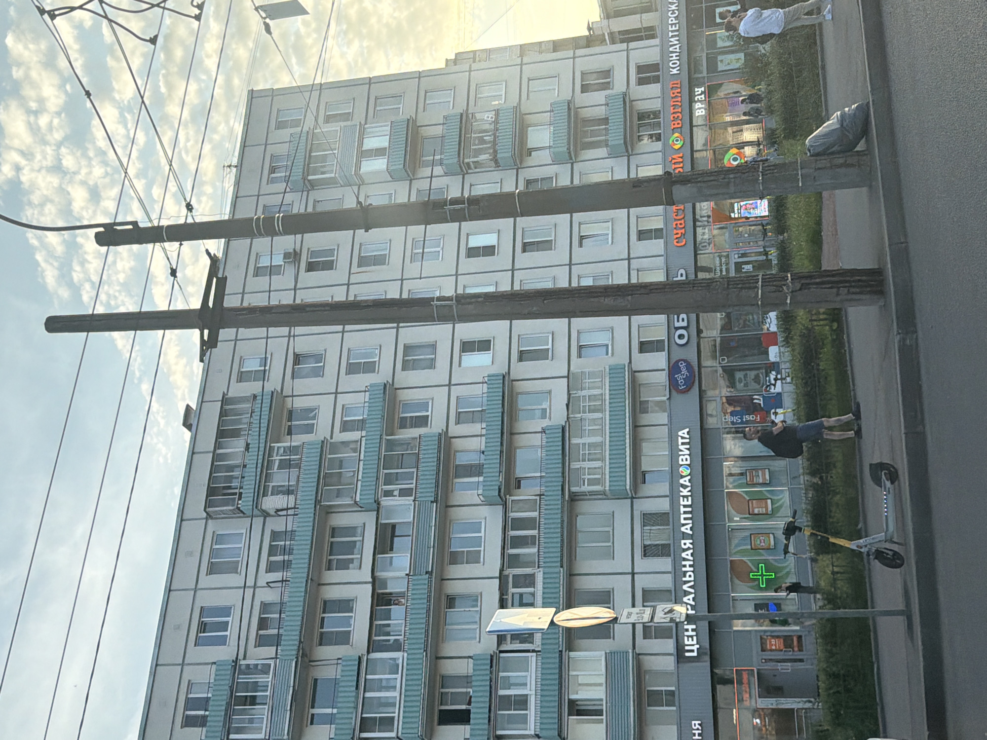 The image depicts a street scene in Saint Petersburg, Russia, with a large, multi-story apartment building dominating the background. The building has a light-colored facade with teal-colored balconies and window frames. In the foreground, a sidewalk runs parallel to the street, with several power poles and overhead wires.
On the sidewalk, a man wearing a black t-shirt and blue shorts is walking away from the camera, carrying a black backpack. A light-colored electric scooter is parked on the sidewalk in front of a storefront. Another person, a woman in dark clothing, is visible through the storefront window, holding a bag.
The storefronts below the apartment building appear to be businesses. One prominent sign reads "ЦЕНТРАЛЬНАЯ АПТЕКАВИТА" (Central PharmacyVita) in large, illuminated letters. Other visible signs include "Fast Step" and what appears to be a confectionery shop with the Cyrillic word "счастье" (happiness) visible. A sign with a green cross, commonly associated with pharmacies, is visible in one of the storefronts.
A large, gray trash bag is leaning against one of the power poles on the sidewalk. The sky is partly cloudy, suggesting it is daytime. The overall scene appears to be a typical urban environment.