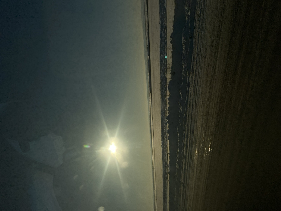The image depicts a vast, snow-covered winter landscape in Озерный, Russia, viewed from a slightly elevated perspective, likely from inside a vehicle.
**Foreground**: The lower third of the frame is occupied by a dark, textured surface, possibly a road or a frozen body of water, showing longitudinal streaks and reflective patches consistent with melting ice or water. Visible reflections suggest the image was captured through a window.
**Middle Ground**: Immediately beyond the foreground, a substantial bank of uneven, piled snow runs horizontally across the scene. Behind this snow bank, an expansive, flat area is uniformly covered in snow, stretching into the distance. Sparse, faint vertical elements, possibly fence posts or distant structures, are discernible across this snow-covered plain.
**Background**: The snow-covered plain extends to the horizon, where a hazy, dark line of distant hills or low mountains is visible.
**Sky**: The sky transitions from a bright, hazy white-blue around the sun to a darker blue-grey towards the top of the frame.
**Sun**: A prominent, intensely bright sun, positioned in the upper-left quadrant, emits strong starburst light rays and exhibits significant lens flare, including a distinct green-blue artifact. Its low position indicates either early morning or late afternoon.
No people, animals, or active subjects are visible. The environment is cold, clear, and rural or semi-rural, characterized by extensive snow cover and bright, low-angle sunlight.