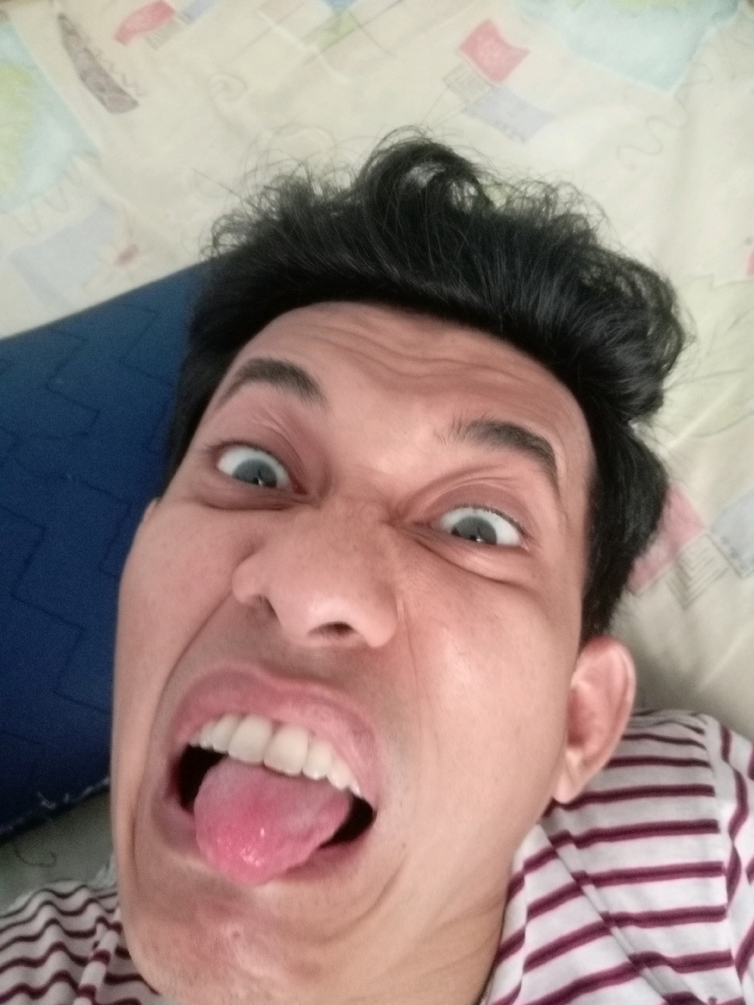 The image shows a close-up, distorted selfie of a person with dark, messy hair. The person is sticking out their pink tongue and has wide, crossed eyes, indicating a playful or silly expression. They are wearing a horizontally striped shirt with maroon and white bands.
The background appears to be a bed with a patterned comforter or sheet in muted colors and a dark blue pillow visible on the left. The lighting suggests it's daytime, possibly indoors. The overall impression is casual and lighthearted, likely taken at home. There is no visible text in the image. The scene is set in Batam, Indonesia.