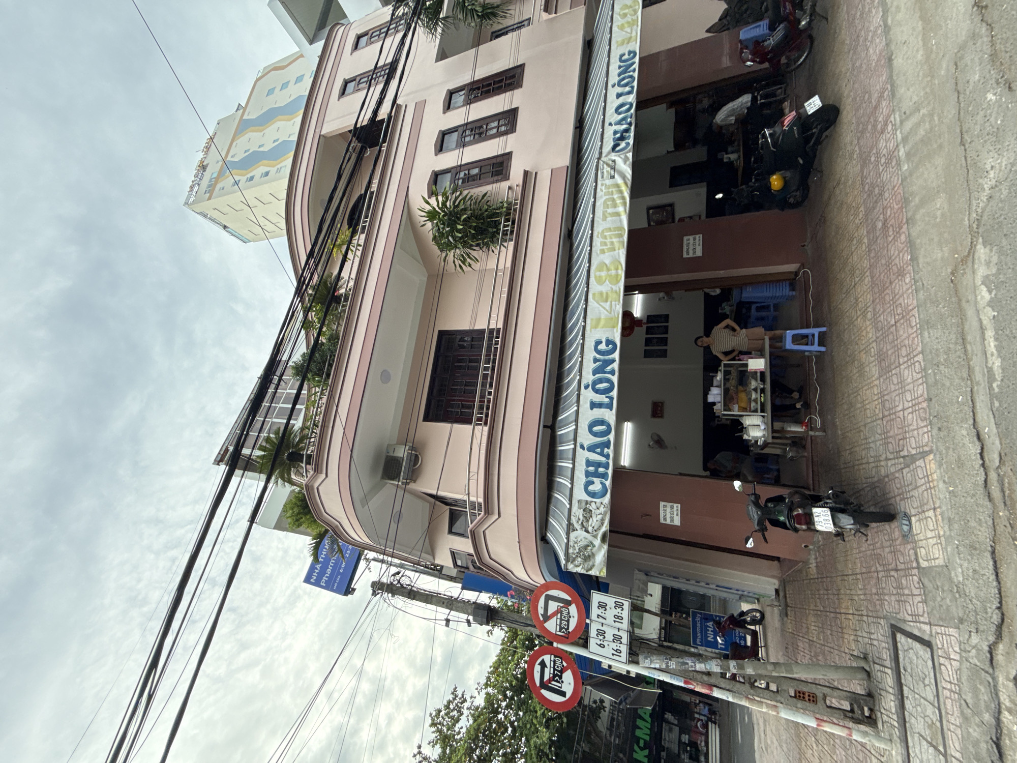 The image depicts a street-level view of a pink building housing a restaurant named "CHÁO LÒNG" at 148 Võ Trứ Street in Nha Trang, Vietnam. The restaurant's awning displays the name in large blue letters. Several motorbikes are parked on the sidewalk in front of the establishment.
On the left side of the image, traffic signs are visible, indicating prohibited turns and hours of operation for certain restrictions. A blue sign for "NHÀ THUỐC PHARMA" (pharmacy) is also present.
The building has multiple floors with balconies and windows. Several potted plants are placed on the balconies. Above the pink building, a taller, modern building with a light-colored facade and blue and yellow wavy decorative elements can be seen.
The sky is overcast, suggesting a cloudy day. The sidewalk is paved with decorative tiles. Several people are visible inside and outside the restaurant, some appearing to be customers or staff. One person, a woman wearing a striped shirt and shorts, stands near a table inside the restaurant. Another person, a man in a checkered shirt, is seated at a table further inside. Another man is standing behind a counter.
