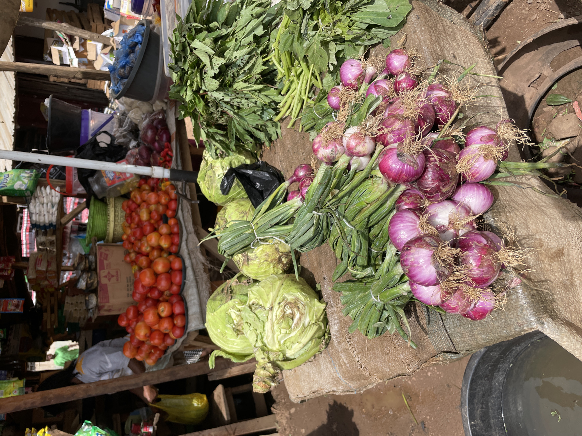 This image depicts a market scene in the city of Mbar, Nigeria. In the foreground, a variety of fresh produce is arranged on a rough, woven mat. Prominently displayed are bunches of red onions with their roots still attached, as well as green onions tied together with string. Behind the onions, several heads of cabbage are visible, some showing signs of wilting. To the right, a large pile of leafy green vegetables, likely spinach or a similar local green, is stacked high.
In the middle ground, a display of ripe red tomatoes is arranged on shallow trays. Further back, several large black buckets and plastic containers are visible. A person wearing a white shirt with yellow trim and dark hair is partially visible on the left, holding a bright yellow plastic bag. In the background, the market structures are evident, with shelves holding various goods and products. The overall environment appears to be an outdoor market with a dusty or dirt ground.