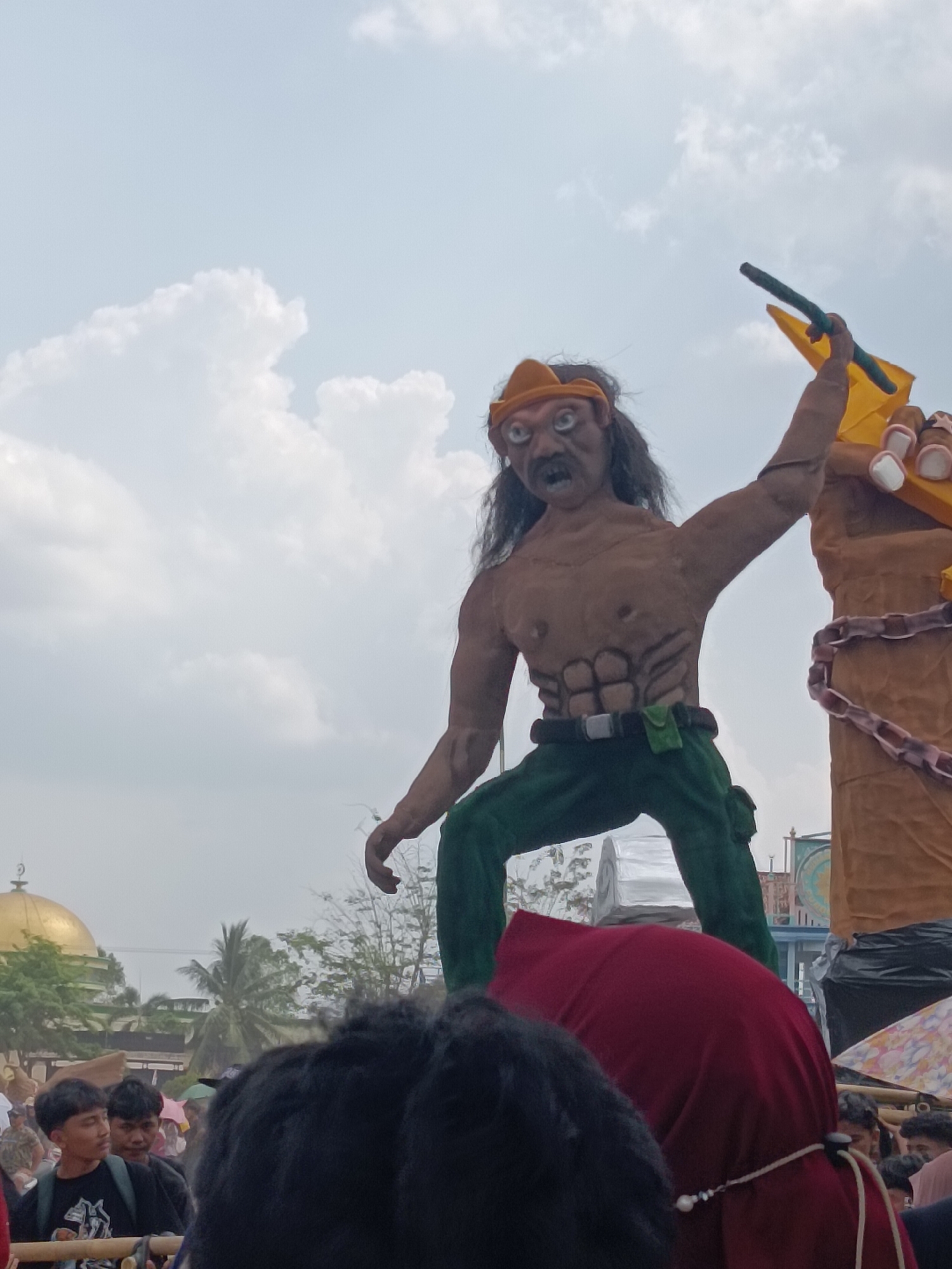 A low-angle, full shot captures a large effigy of a muscular, tan-skinned man wearing a yellow bandana, a brown-and-green patterned shirt, and green pants. The effigy has a sinister facial expression, with wide, white eyes and a gaping mouth. It holds a green stick aloft in its right hand. The effigy stands in front of a cloudy sky, with a golden-domed building and palm trees visible in the background to the left. The bottom of the image shows a crowd of people, some wearing black shirts, and parts of red and black clothing. The scene appears to be an outdoor gathering or festival. The effigy is likely part of a religious or cultural event, possibly a parade, in Tegal Gondo, Indonesia.