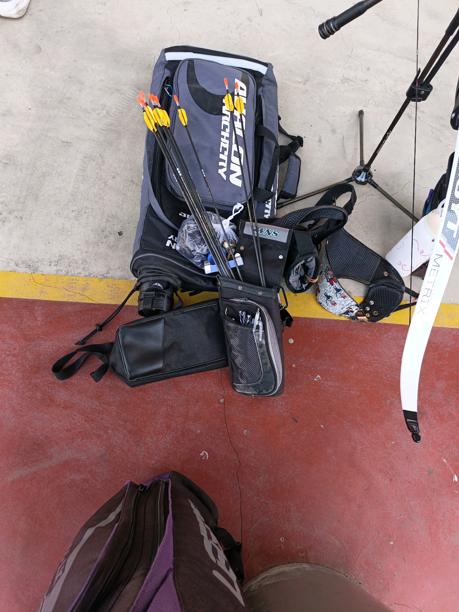 In Jonggrangan, Indonesia, an archery equipment setup is visible on a concrete surface bordering a red-painted area. The central focus is a large, gray archery bag with "ARCHERY" printed vertically in white. Several arrows with yellow fletching and orange nocks are inserted into the bag. Adjacent to the bag is a quiver containing pens and what appear to be other small archery accessories. A black pouch with a zippered compartment lies to the left of the bag. A white riser with "METRIX" printed in black and red lettering is positioned on the right. The scene also includes a black archery stand with folded legs and a black stabilizer extending upwards. A black backpack with purple accents is partially visible in the lower left corner of the frame. The ground is a mix of concrete and red paint, with some debris scattered on the surface.
