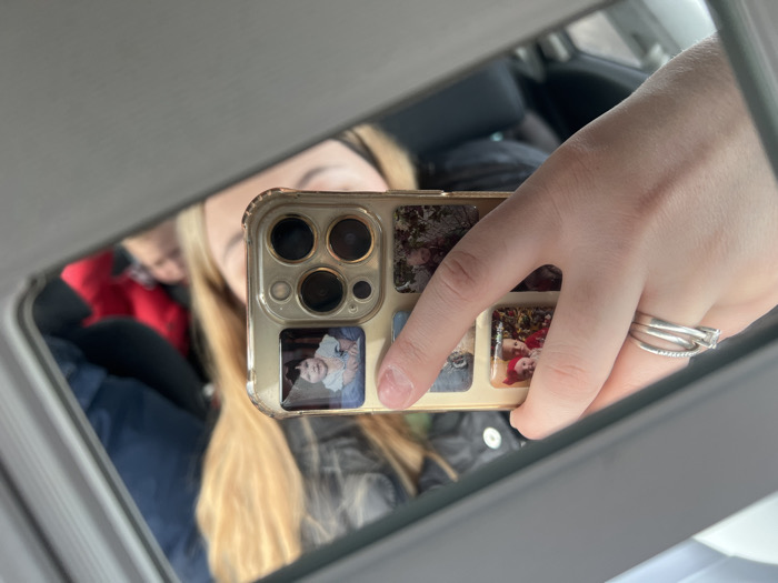 The image displays a reflection within a vehicle in Chaltyr', Russia. The central subject is a light-skinned right hand, wearing two rings on the ring finger (one a thin band with small stones, the other a thicker, twisted band featuring a prominent clear stone), holding a gold-colored smartphone. The smartphone case exhibits a triple-lens camera array and is customized with multiple small photographs on its rear, including a clear image of one young child and another of two young children. The individual holding the phone is partially visible in the reflection, showing long, blonde hair and dark clothing. Behind this individual, a second person in red attire is faintly reflected in the vehicle's interior. The surrounding environment consists of grey car interior elements, indicating a vehicle cabin during daytime.