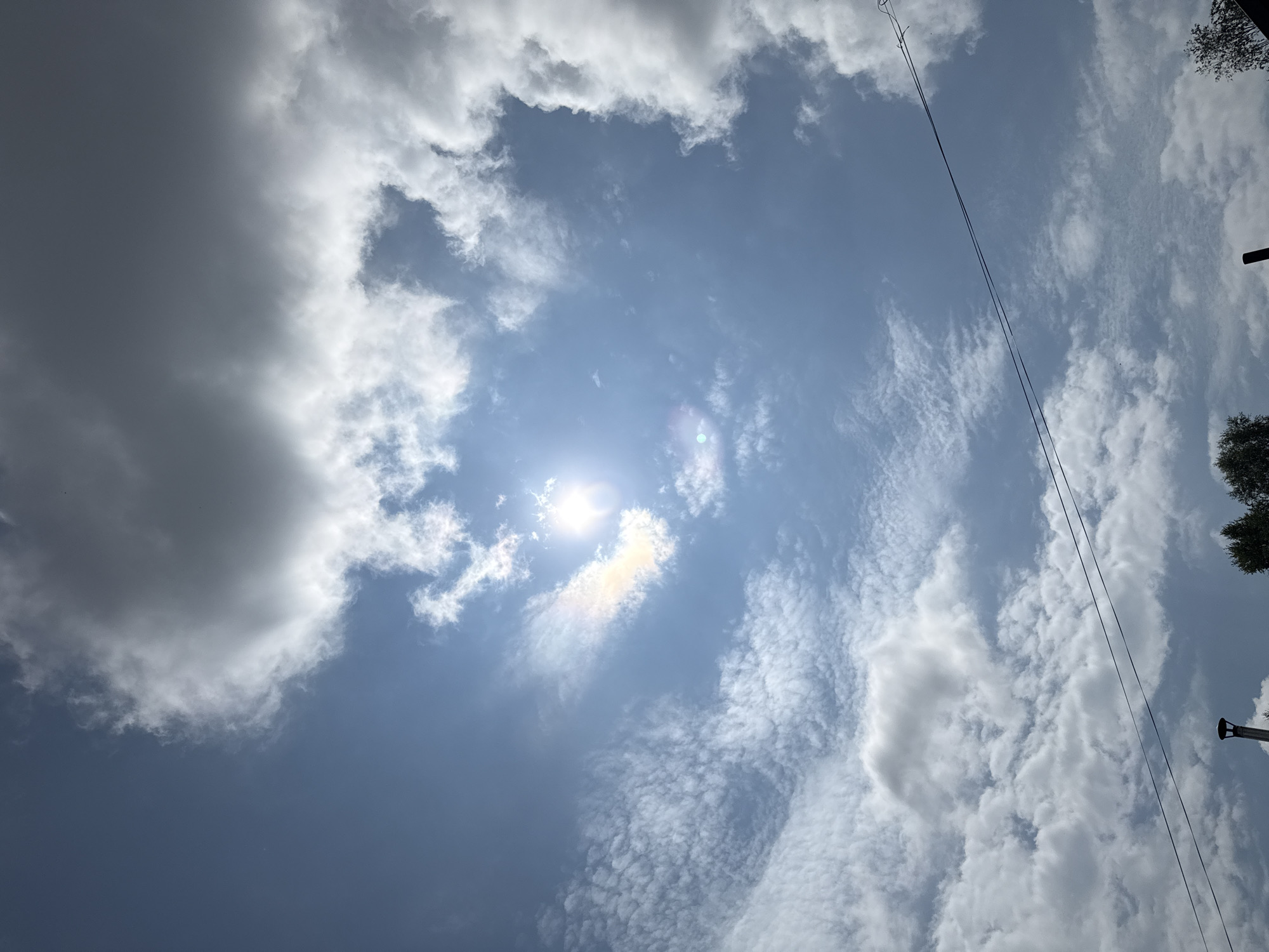 The image shows a bright, partly cloudy sky. The sun is visible and shining through a gap in the clouds, creating a hazy glow with a faint rainbow effect around it. Some clouds are dark and heavy, while others are wispy and white. Two dark electrical wires are strung diagonally across the lower portion of the sky. In the very bottom of the frame, silhouetted against the sky, are the tops of some trees and what appears to be a chimney or ventilation pipe. The scene is likely outdoors and appears to be a daytime view from a residential area. The location is given as Krasnogorskoye, Russia.