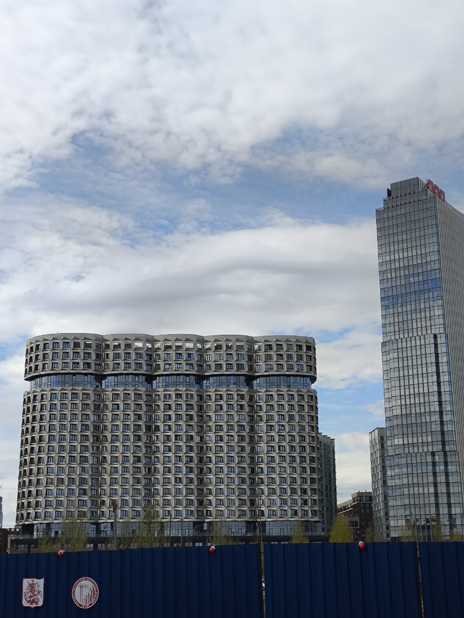 The image shows two modern buildings in Moscow, Russia, under a partly cloudy sky. In the foreground is a blue metal fence with two circular emblems. Behind the fence, the building on the left has a unique, curved design with multiple floors and small windows. The building on the right is a tall, rectangular skyscraper with a grid-like facade and a red logo at the top. Some trees are visible between the fence and the building on the left. The overall scene suggests an urban environment with contemporary architecture.