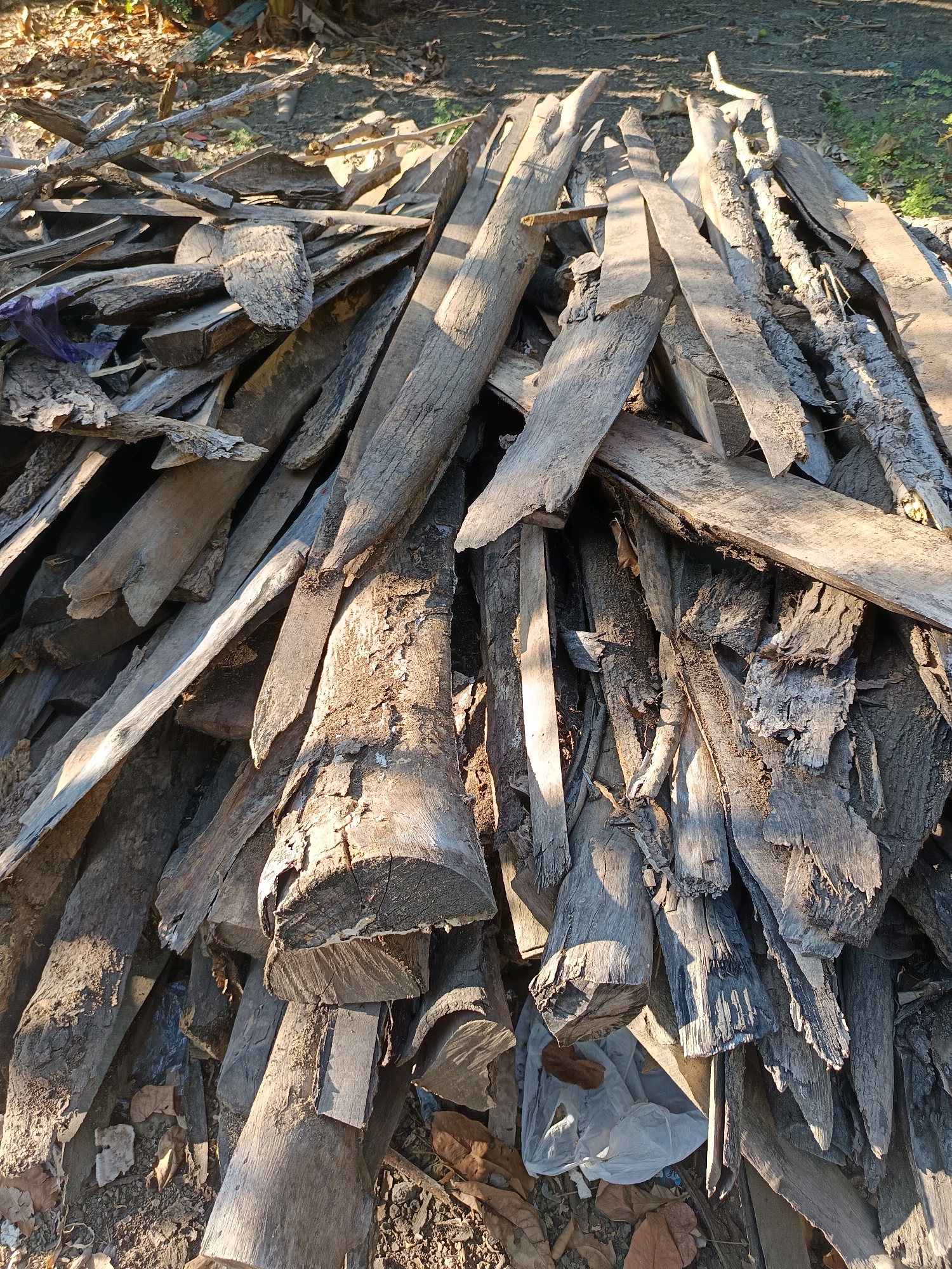 The image depicts a large, haphazard pile of weathered, gray-toned wooden debris. The wood varies in size and shape, consisting of planks, logs, and irregularly broken pieces, some appearing to be salvaged or discarded. The texture of the wood is rough and aged, with visible grain and splintered edges. Sunlight casts strong shadows across the pile, highlighting the uneven surfaces and creating a chiaroscuro effect. The background shows a ground surface covered with dry leaves and some sparse vegetation, suggesting an outdoor environment. There are no people or animals visible in the image, and no specific actions are being depicted. The overall impression is one of disarray and the accumulation of natural materials. The location is specified as Gunung Sekar, Indonesia, an area known for its natural landscapes and cultural significance.