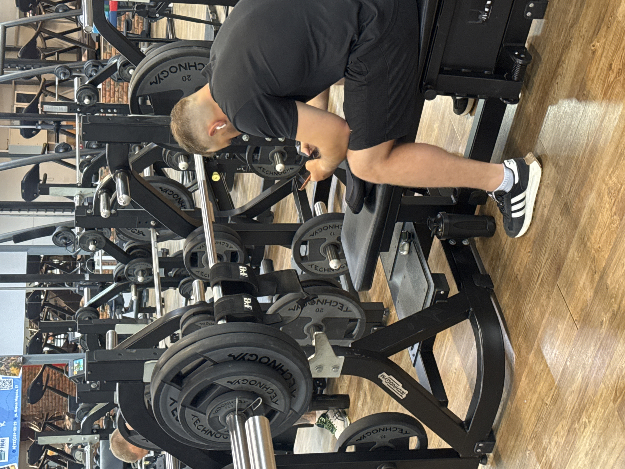 The image, oriented sideways, captures an indoor gym scene in Chaikovsky, Russia. The primary subject is a male individual with short, light-colored hair, wearing a black t-shirt, black shorts, white socks, and black Adidas sneakers with white stripes. He has white wireless earphones in his ears. He is bent over, facing downwards, seemingly interacting with or resting on a black weightlifting machine, possibly adjusting settings or weights.
The setting is well-equipped with numerous black Technogym brand machines and weights. Various circular weight plates, marked "TECHNOGYM" and displaying values like "10" and "20", are visible on the barbells and machine racks. The floor is made of light-colored wood. In the background, more gym equipment is discernible, suggesting a spacious fitness facility. A small detail shows "D.F" on a strap attached to one of the weight machines. A poster in the background, although largely obscured, features text including "PEGAS", a phone number "8(922)389-12-29", and an address "ул. Карла Маркса, 27", which confirms the location in Russia. No specific time of day or emotional state is clearly evident.
