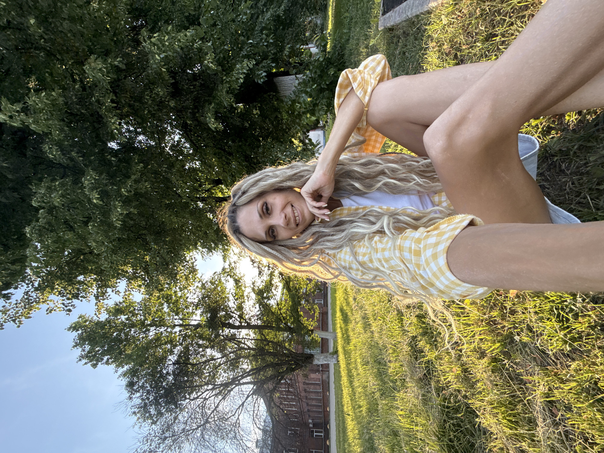 In an outdoor setting, likely a park or grassy area within the city of Shebunino, Russia, a young woman with blonde dreadlocks is seated on the grass. She is wearing a yellow and white checkered shirt over a white top and denim shorts. The sun is setting, casting a warm glow and long shadows across the lawn. In the background, a brick building with multiple windows and trees are visible. The woman is looking at the camera and smiling, with her hand resting on her chin. The image captures a moment of leisure in a suburban or semi-rural environment.