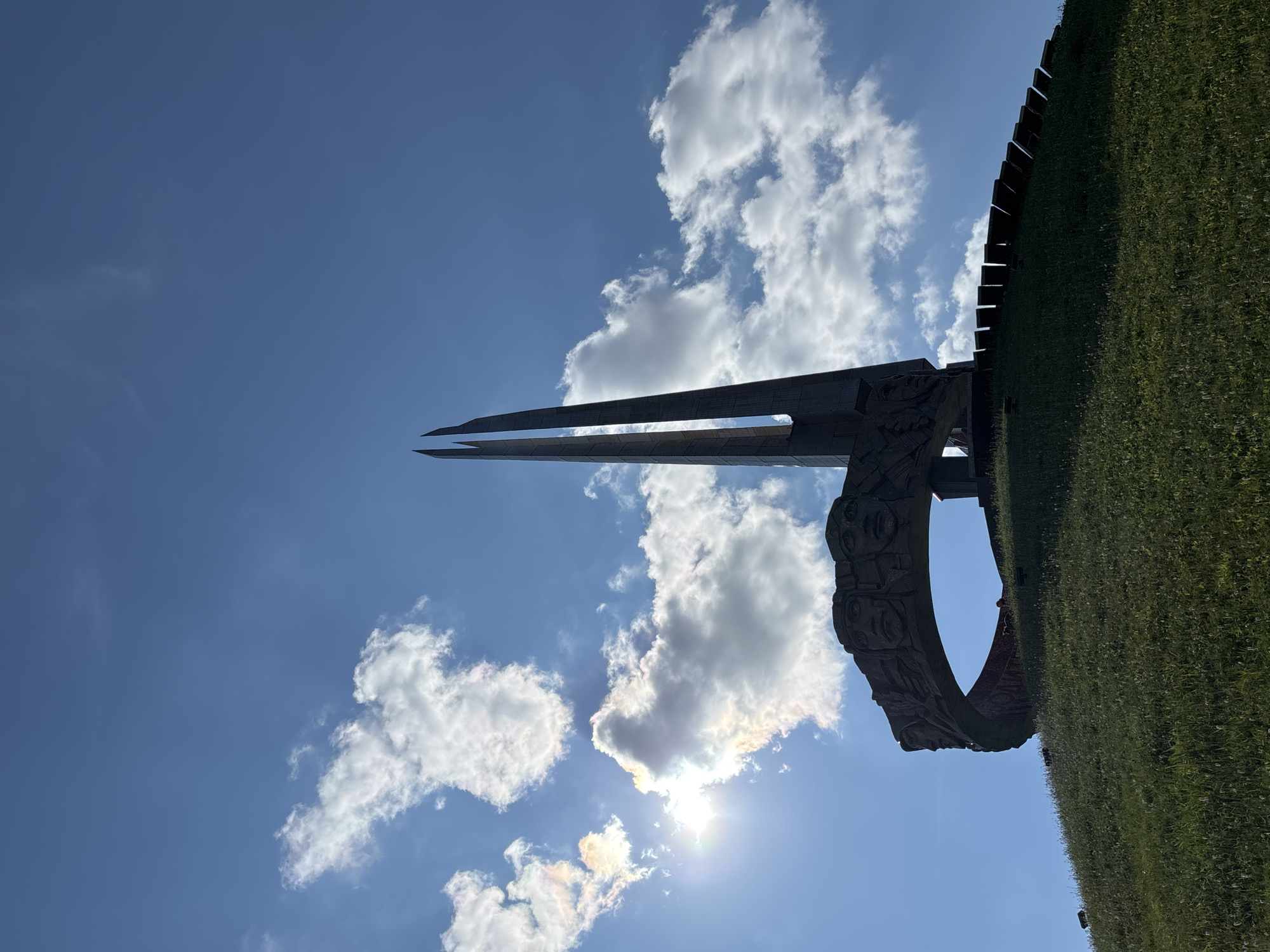 A wide shot captures a monumental structure against a bright blue sky with scattered white clouds. The monument is silhouetted against the sun, which shines with a slight halo effect. The monument features a large, circular stone ring adorned with carved faces, resting atop a grassy hill. Two tall, pointed spires rise from behind the ring. The hill is covered in green grass, and a series of dark, rectangular elements are visible along its upper edge. The overall impression is one of solemnity and grandeur, with the monument standing as a prominent landmark in the landscape.