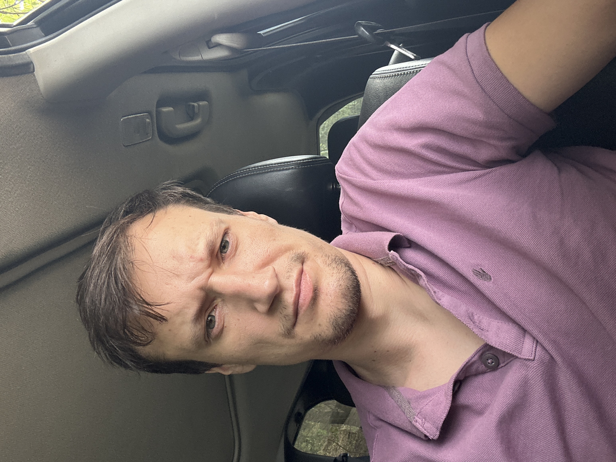 The image is a selfie taken inside a car. A man with short, dark brown hair and a light beard is looking at the camera with a furrowed brow and a slightly crooked mouth, suggesting a neutral or slightly displeased expression. He is wearing a light purple polo shirt with a two-button placket. The car's interior is visible, including the black leather seat he is sitting in and the car's headliner above him, which appears to be a dark gray material. Natural light is coming from the right side of the frame, illuminating the scene. The specific location in Makiv, Ukraine cannot be determined from the visual information provided in the image.