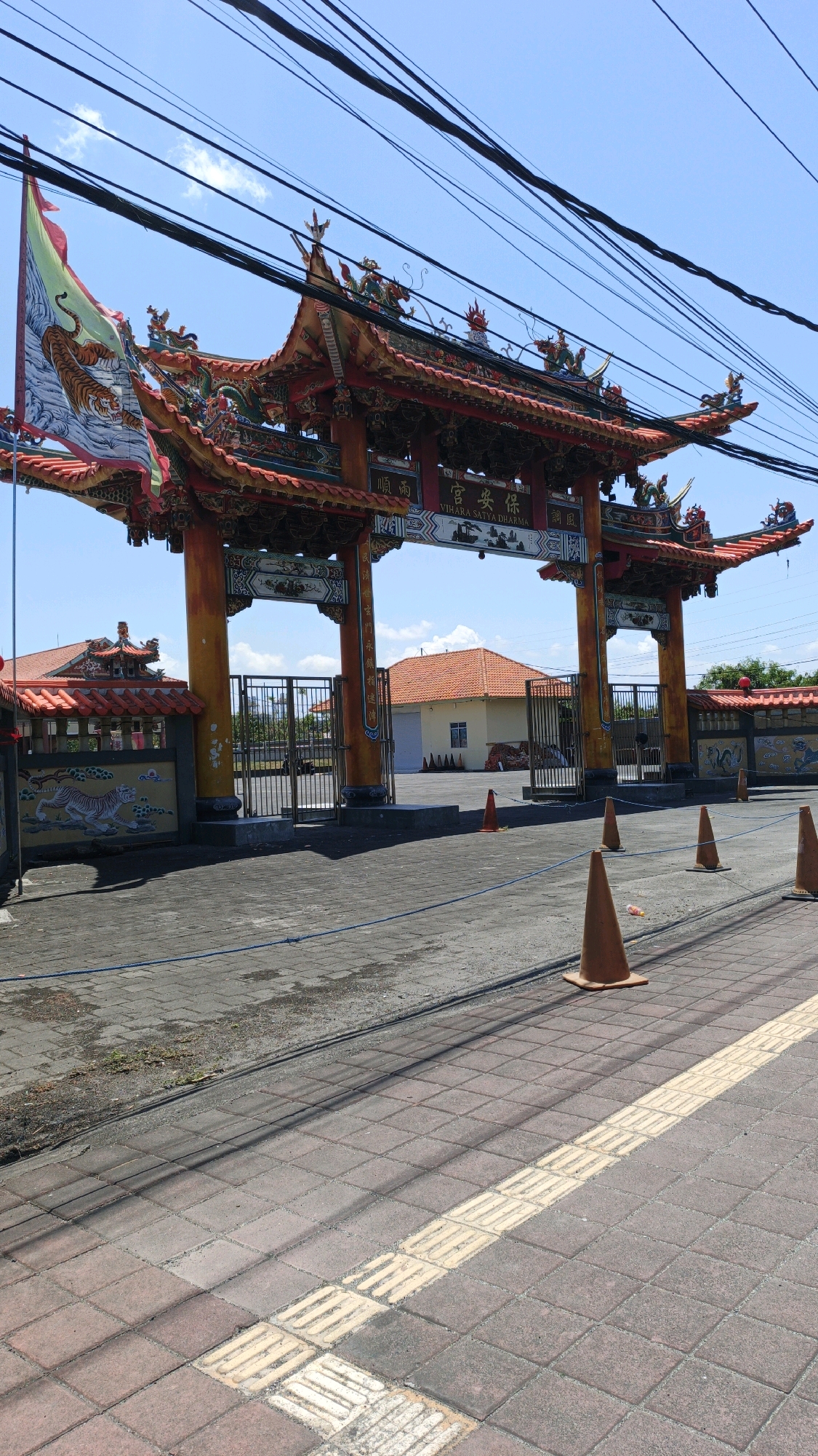 The image shows the entrance to Vihara Satya Dharma, a Buddhist temple located in Denpasar, Indonesia. The entrance is a large, ornate gate with a red and gold roof, decorated with dragons and other mythical creatures. A flag with a tiger image is flying on the left side of the gate. Behind the gate, there are metal fences and buildings with orange tile roofs. The ground in front of the gate is paved with bricks and asphalt, and there are several orange traffic cones placed along the road. Power lines are visible in the sky, casting shadows on the ground. The sky is clear and blue with a few clouds. The scene appears to be taken during the daytime, under bright sunlight.