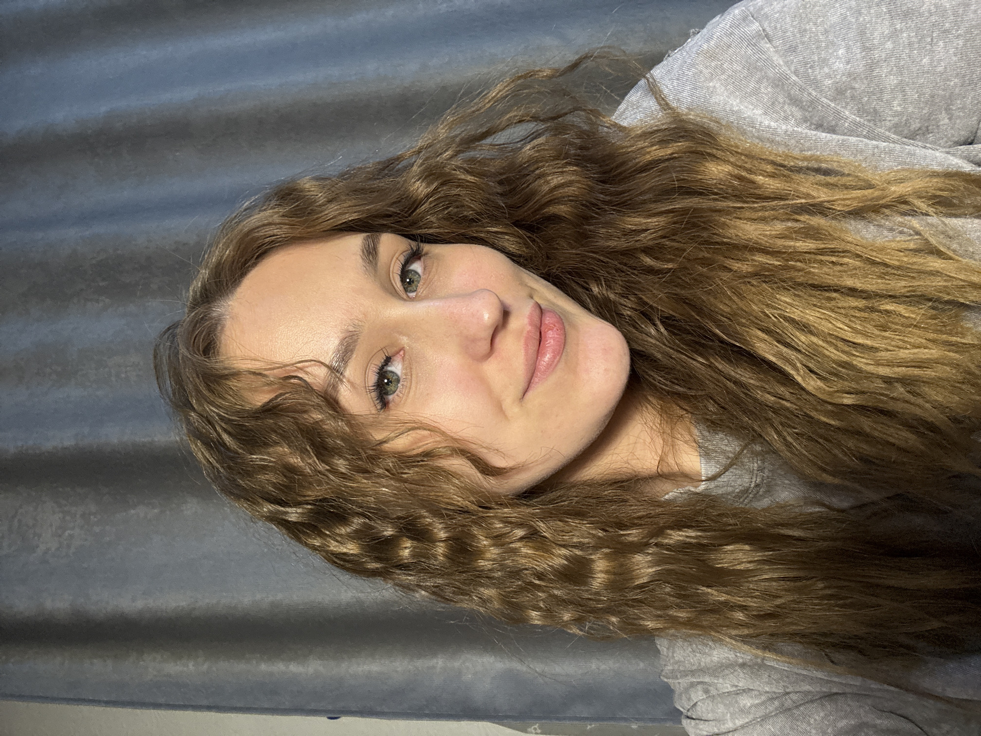 A woman with long, curly brown hair is centered in the image. She is wearing a grey shirt. She has green eyes and a neutral expression. The background is a grey curtain. The location cannot be determined to be in Марковский, Russia, from the given image.