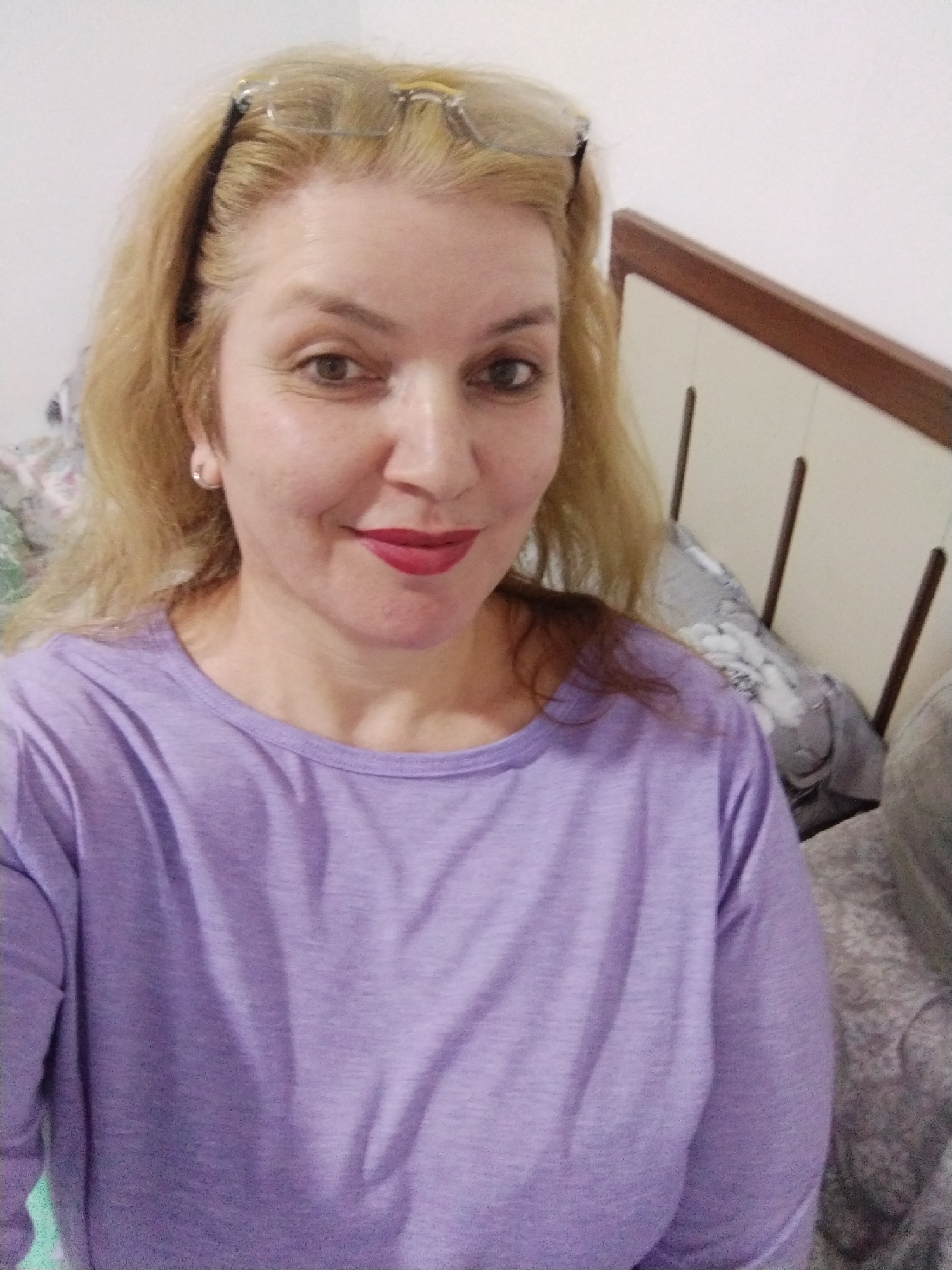 The image shows a middle-aged woman with blonde hair wearing glasses on her head and a purple long-sleeved shirt. She is wearing lipstick. The background includes a bed with a headboard and patterned pillows. The location is likely indoors, but not identifiable within Al Mas'oudi, United Arab Emirates.