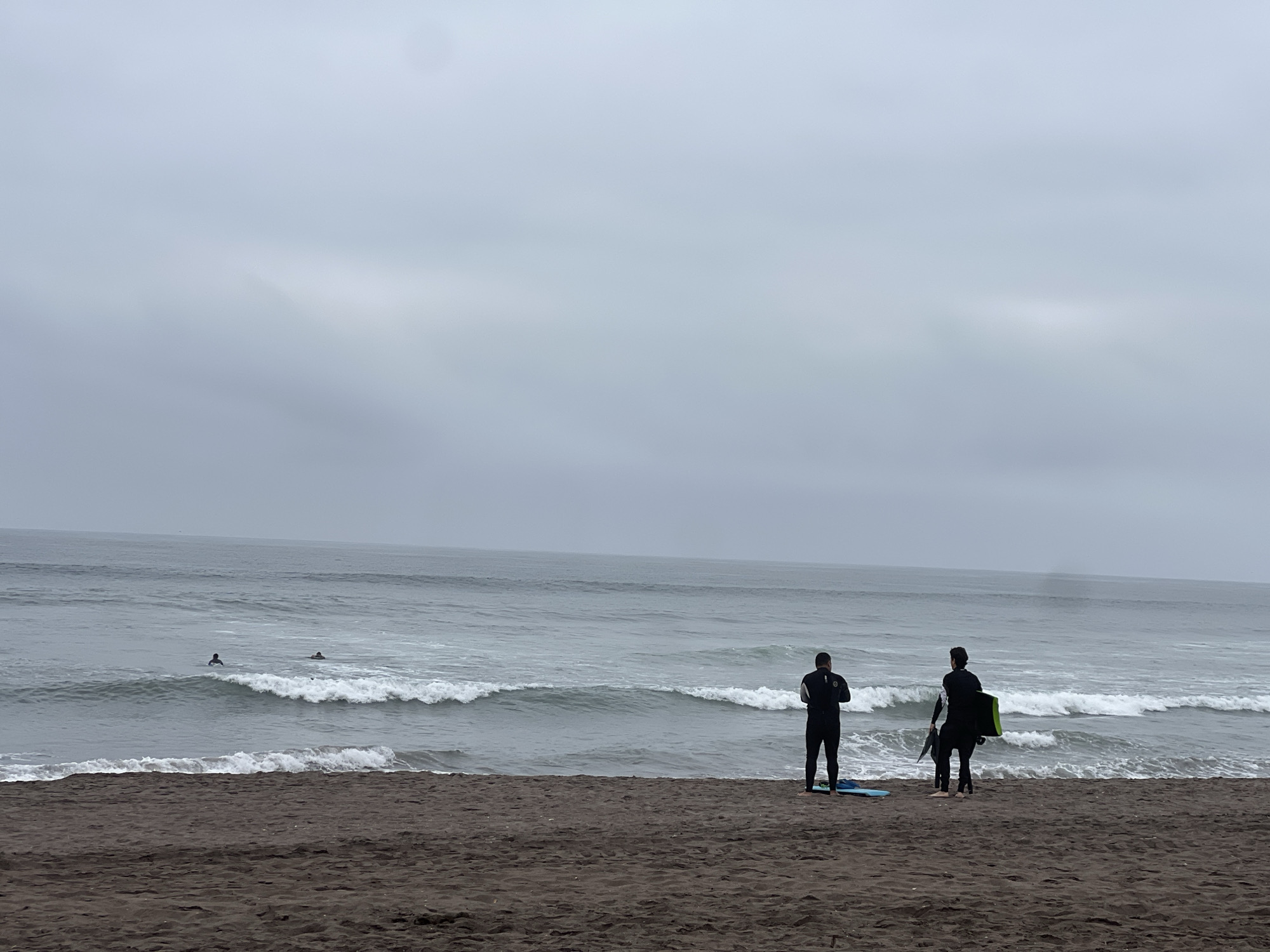 Two surfers in black wetsuits stand on a dark sand beach, facing the ocean under a cloudy sky. One surfer on the right holds a bright green bodyboard. Further out in the gray ocean, two other figures are visible, one appearing to be on a surfboard and another floating. Waves are breaking on the shore. The scene is likely La Lisera Beach in Arica, Chile, known for its dark sand and frequent surfing activity, especially on overcast days.