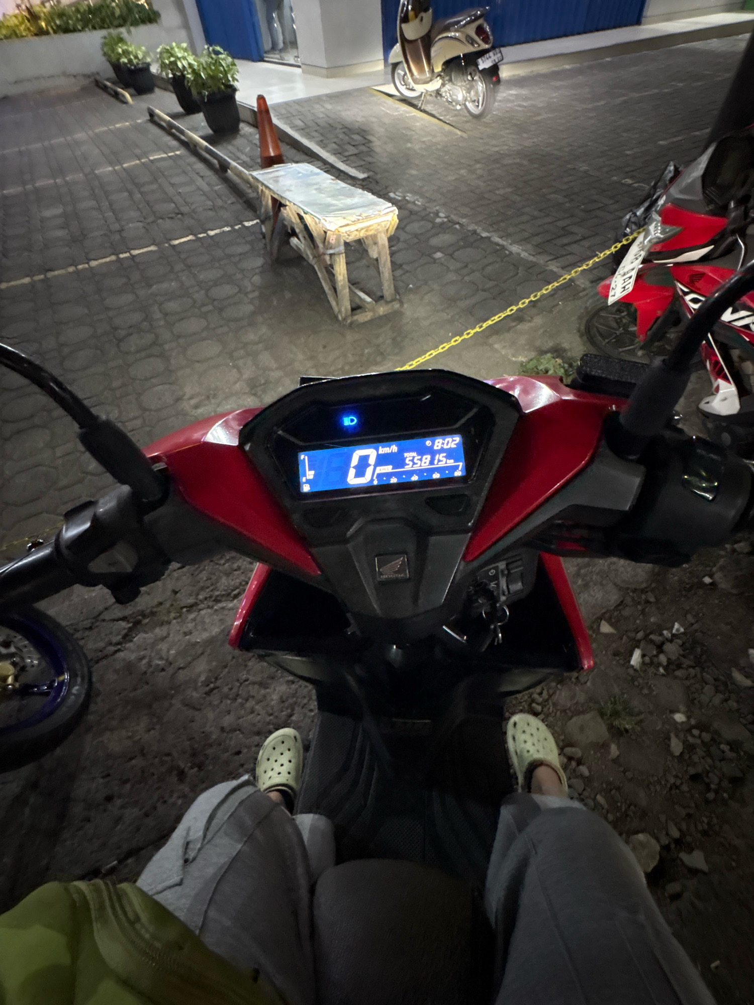 The image depicts a first-person perspective of someone sitting on a red motorcycle at night in a paved outdoor area. The motorcycle's digital dashboard is visible, showing a speed of 0 km/h and a time of 8:02. A portion of a red and white motorcycle is parked to the right. In the background, there is a cream-colored scooter parked under a building with blue walls. The ground is paved with interlocking bricks, and there are several potted plants and a construction barrier with orange cones in the distance. The scene appears to be a parking lot or street area. The presence of the red and white motorcycle with a license plate ending in '27' and the cream-colored scooter suggests a common urban setting. While the exact location is not specified, the details observed are consistent with a public space in a city like Bandung, Indonesia.