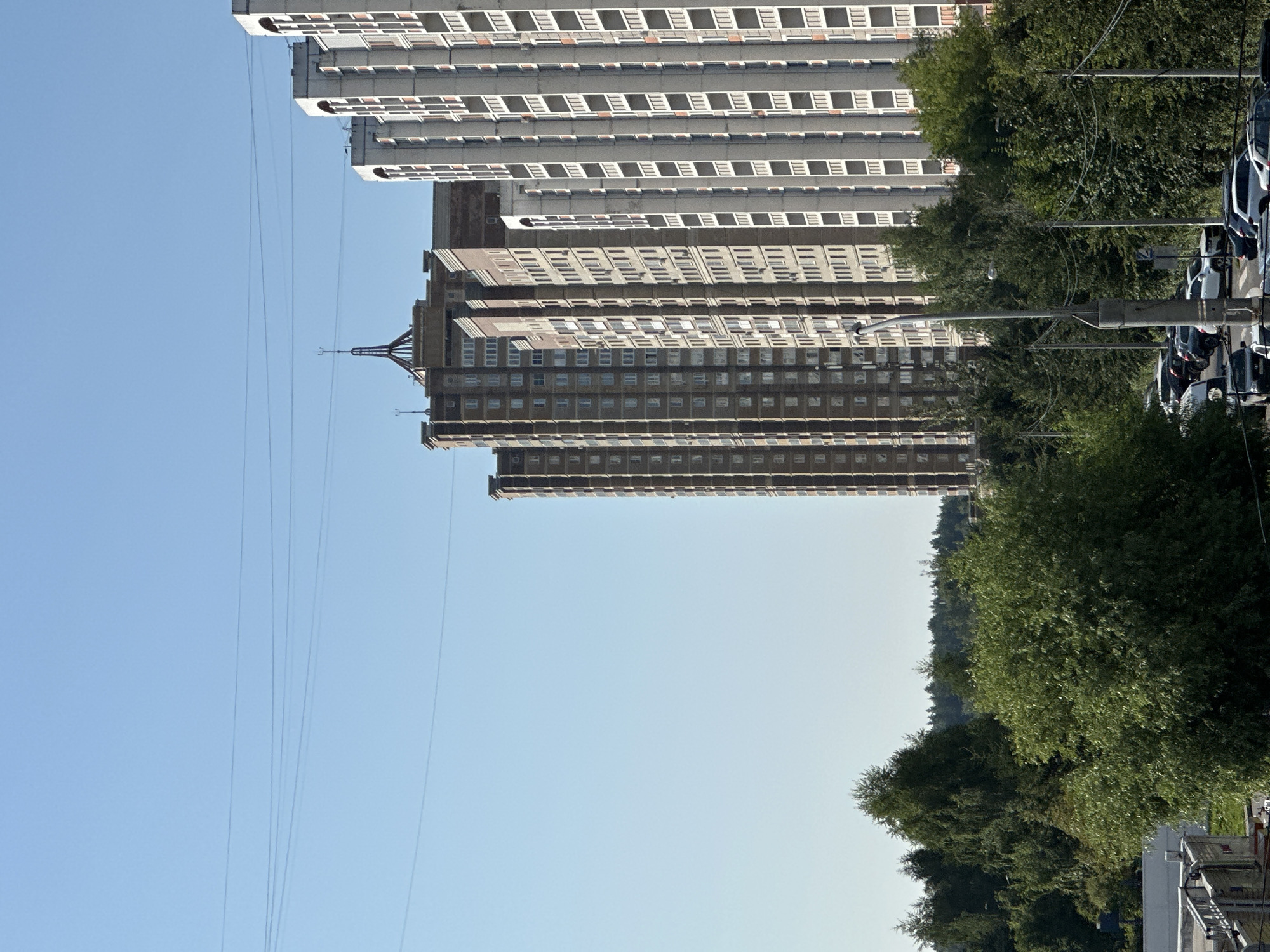 The image shows a view of apartment buildings in Podolsk, Russia, on a clear day. Several tall, multi-story apartment buildings are visible, with some appearing to be under construction or renovation due to exposed concrete and unfinished balconies. The buildings are arranged in a row, with some older, darker brick buildings and newer, lighter-colored structures. Trees and greenery are present in the foreground and between the buildings, and a line of power lines stretches across the sky. A street with parked cars is visible at the bottom of the image, suggesting an urban or suburban setting. The scene is static, with no people or animals clearly depicted.