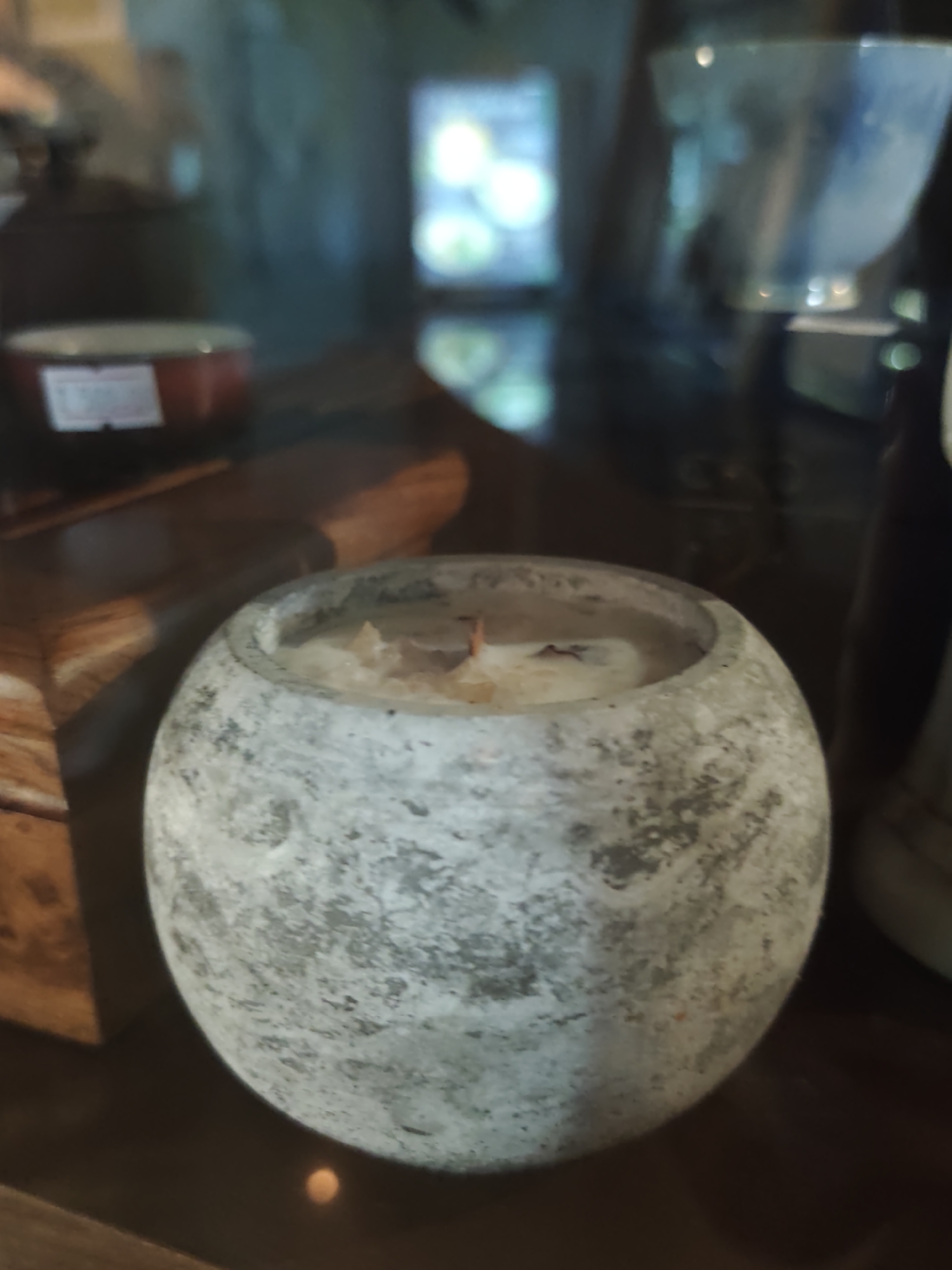The image shows a close-up of a round, textured, gray candle in a concrete holder. The candle appears to be lit, with a small flame visible at its center. The holder has a rough, mottled surface. In the background, out of focus, are various objects on a wooden surface, including a red dish with a white label and a blurry blue and white bowl. Further in the background, a brightly lit object with circular lights is visible, possibly a display or screen. The overall setting appears to be indoors, possibly a shop or cafe. The image does not contain information to determine its location in Sochi, Russia.