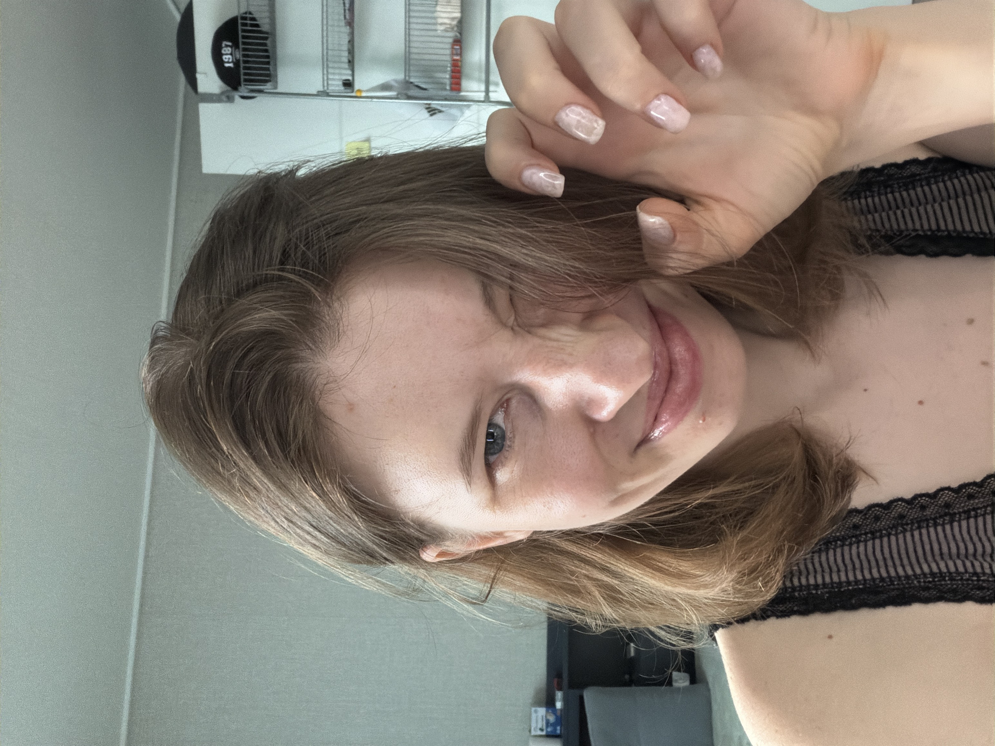 A young woman with light brown hair and blue eyes is winking at the camera. She is wearing a black lace top. Her right hand is raised to her face, with her fingers splayed. Her nails are painted a light, pearlescent color. The background appears to be an indoor setting, possibly a bedroom or living area, with a wall and shelves visible. A black baseball cap with the year "1987" printed on it is seen on one of the shelves. The lighting suggests it is daytime. There are no explicit indicators of the specific location within Dongducheon-si, South Korea.