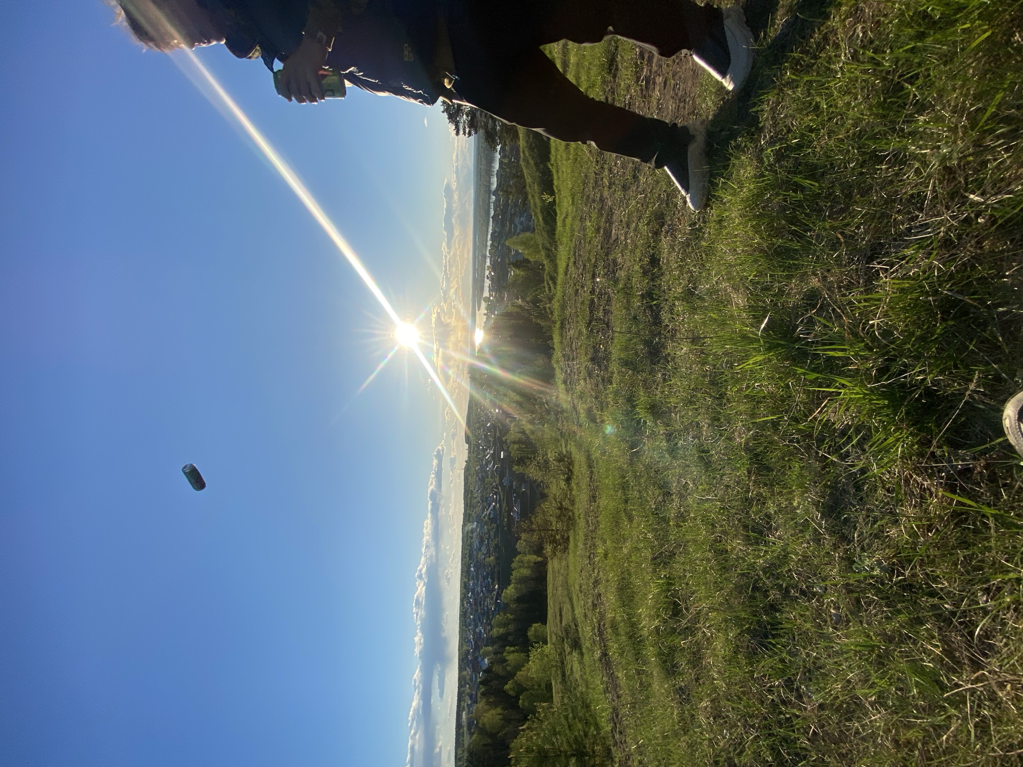 A person stands on a grassy hill in Ольховка, Russia, overlooking a cityscape under a bright sun. A can is in the air, with the person possibly having thrown it. The city extends to the horizon with a river or large body of water visible.