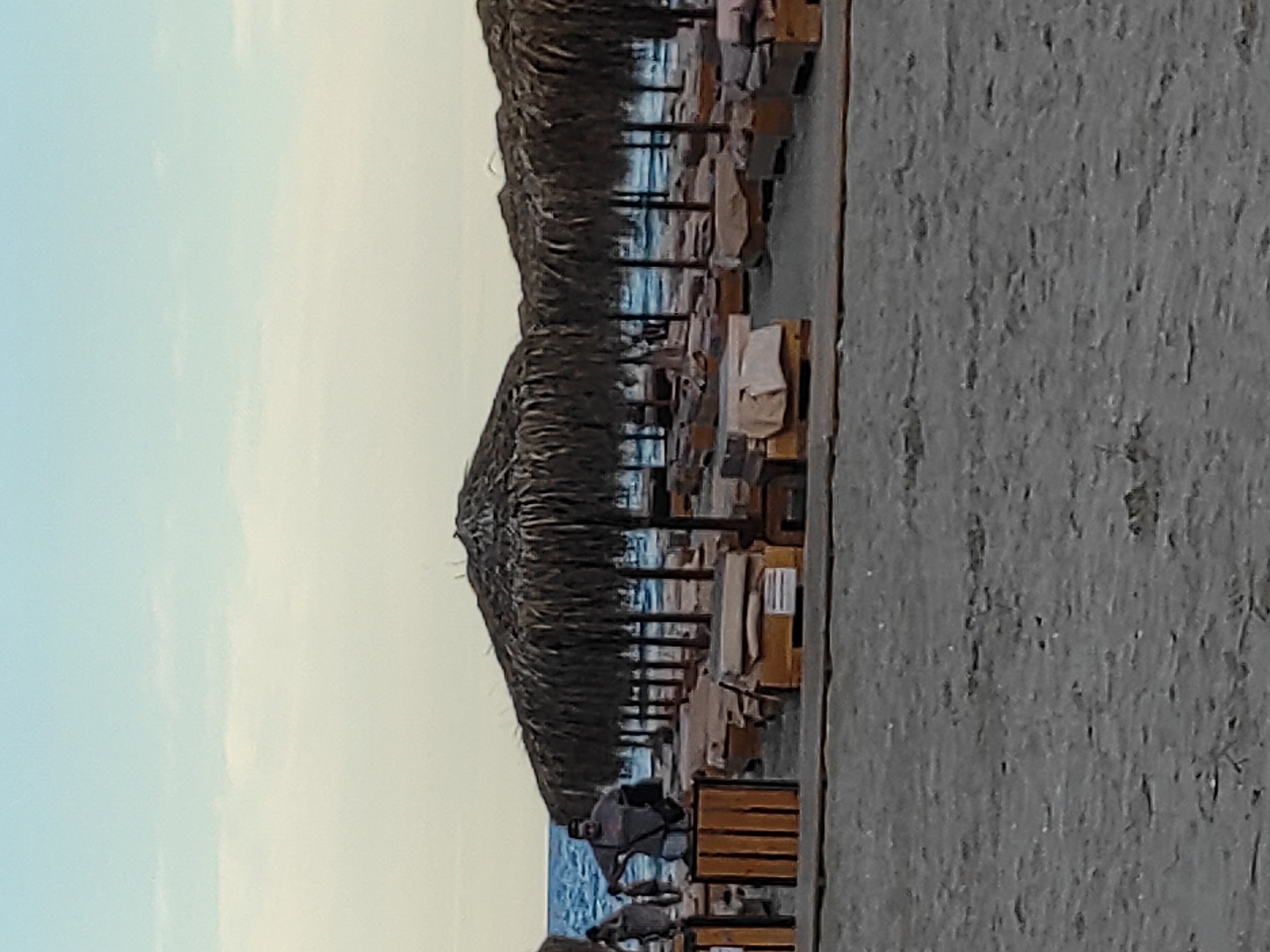 The image depicts a beach scene at Mamaia Nord, Năvodari, Romania, likely during daylight hours given the bright sky. In the foreground, the sandy beach is visible with textured patterns. Beyond the sand, a row of thatched umbrellas shades empty lounge chairs. The chairs are arranged in neat rows, some covered with light-colored fabric. The ocean, a vibrant blue, stretches across the background. Two individuals are present on the left side of the frame, standing near a wooden structure with vertical slats. One person, wearing a dark shirt and lighter shorts, is facing away from the viewer and appears to be interacting with an object in their hands. Another individual is to their left, also facing away. The overall impression is of a beach resort setting.