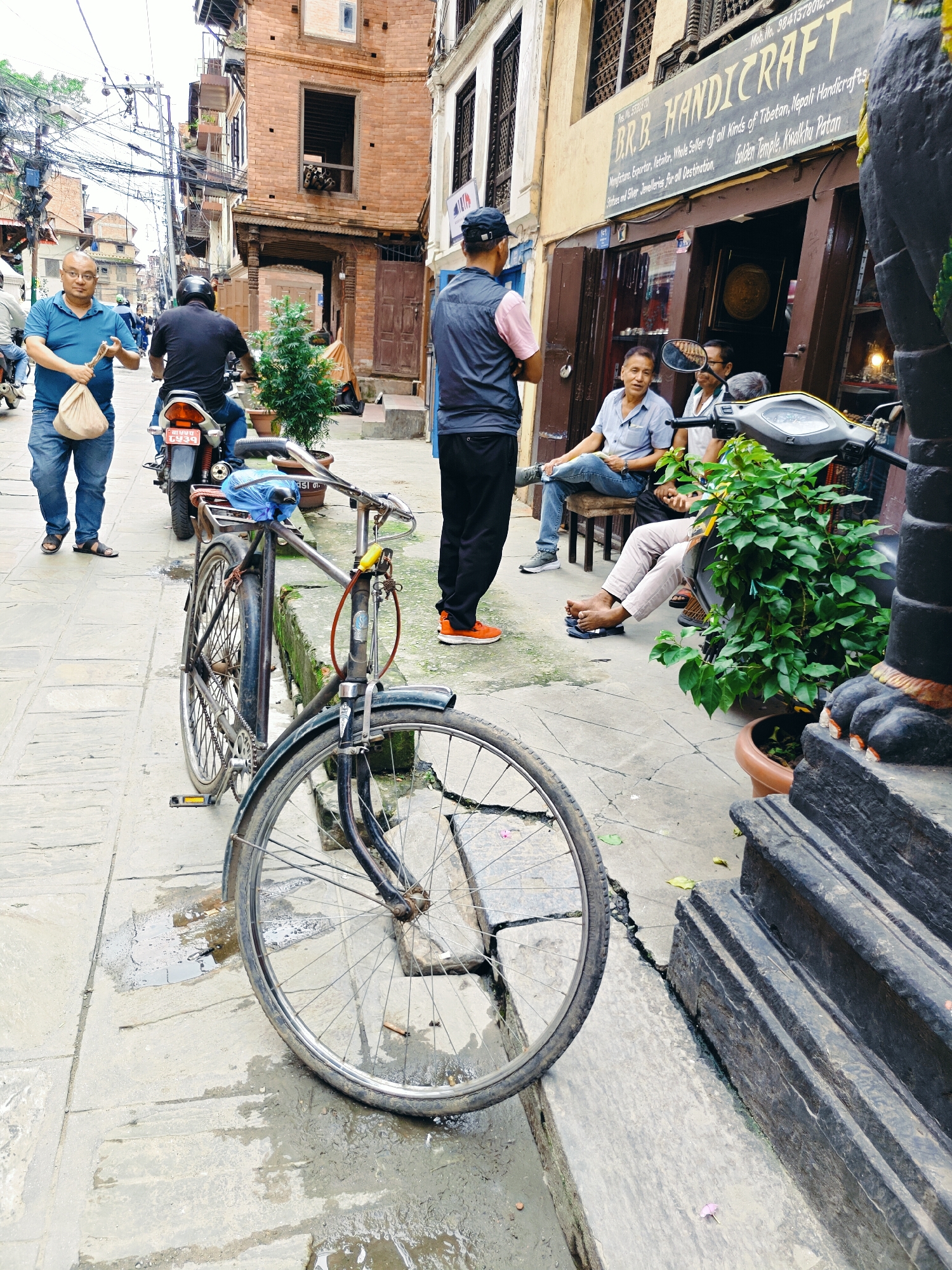 The image depicts a street scene in Lalitpur, Nepal, likely near Patan Durbar Square, given the presence of traditional architecture and handicraft shops. In the foreground, a black bicycle with a blue plastic cover on its seat is parked on a wet, stone-paved sidewalk. The bicycle's front wheel is on the right side of the frame, and the rear wheel is visible on the left.
Several individuals are present in the midground and background. A man in a blue t-shirt and jeans stands on the left, holding a light-colored bag. Behind him, a person wearing a black t-shirt and helmet rides a motorcycle. Further back, another individual in a dark blue vest and a black baseball cap stands facing away from the camera.
To the right, a storefront with the sign "B.R.B. HANDICRAFT" is visible. Three men are seated outside the shop on a low wall or bench. One man in a light blue shirt is holding a book or papers. Another man beside him is wearing a white t-shirt. A third man, wearing a light-colored shirt and pants, is also seated. A motorcycle is parked in front of the shop.
The environment shows traditional brick buildings with wooden accents and balconies. Overhead, numerous electrical wires crisscross the sky. The ground is wet and appears to have puddles, suggesting recent rain. Green potted plants are placed near the storefront and the seated individuals. The overall impression is of a bustling, albeit somewhat damp, street in a historic urban area.