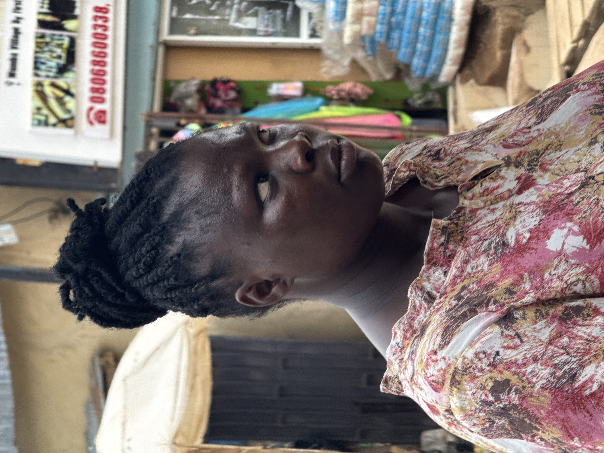 A medium shot shows an African woman with a serious expression, appearing in front of a store in Abuja, Nigeria. She has black braided hair styled in a bun and wears a patterned pink and brown shirt. In the background, the store displays various items under a white awning, with a sign showing the phone number "08068600338". The environment suggests a commercial area within Abuja.