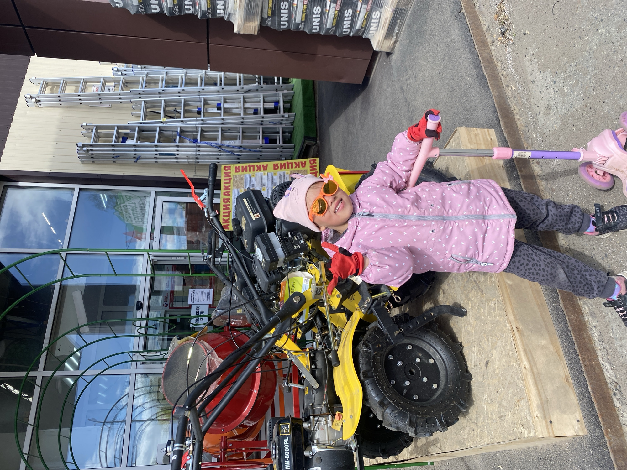 The image shows a girl wearing a pink jacket, a pink hat, and orange sunglasses, lying on a piece of machinery (possibly a two-wheel tractor or cultivator) outside a shop. She is smiling and holding the handles of a pink and purple scooter. Behind her, there's a display of ladders and stacks of UNIS-branded construction materials. The location is likely Chaikovsky, Russia, based on the Cyrillic script on the sale signs and the side of the machinery, and the "MK-8000PL" model number is visible on the machine. The environment appears to be a retail or hardware store's outdoor display area.