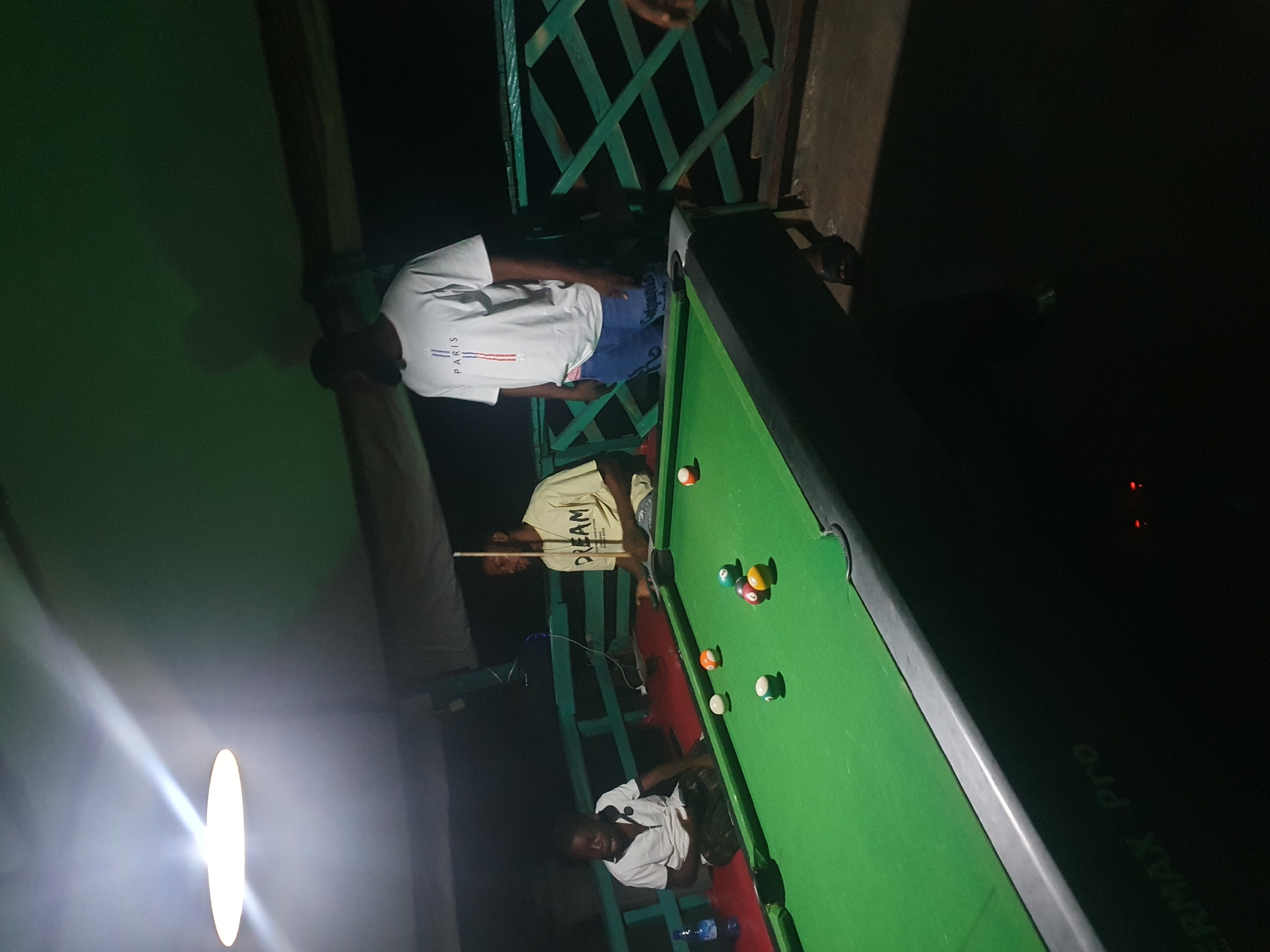 The image shows a group of young men playing pool on a covered outdoor patio in Lokoja, Nigeria, at night. A bright overhead light illuminates the scene. On the left, two men are seated on a red bench near a pool table. One man, wearing a white polo shirt, is looking down at the table. Another man in a white shirt with a black collar is looking at something off-camera. In the center of the image, a man wearing a light yellow t-shirt with the word "DREAM" printed on it is standing, holding a pool cue. To the right, a man in a white t-shirt with "PARIS" written on it is standing near the pool table. The pool table is green with several colored balls on it. The background consists of a dark green, lattice-like railing and dark shadows. The overall atmosphere suggests a casual nighttime gathering for recreation.