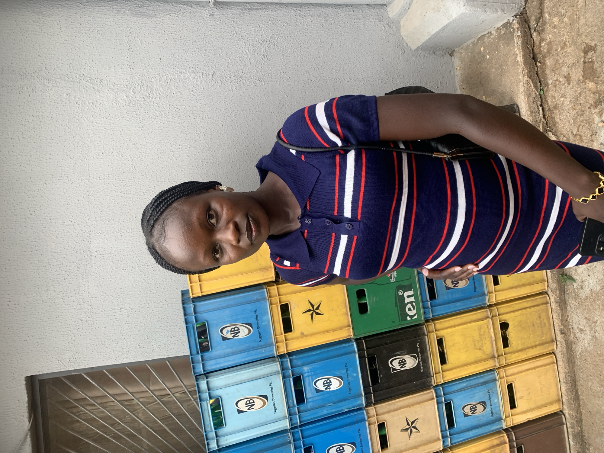 A woman stands in front of a stack of beer crates in Jos, Nigeria. She has dark skin, braided hair, and is wearing a navy blue, red, and white striped dress. She is looking at the camera. She wears a gold bracelet on her left wrist, and she holds a smartphone. Behind her, there is a light gray concrete wall and a window with a metal grill. The beer crates are stacked in a disorganized manner and are various colors, including blue, yellow, green, brown, and black. Some of the crates are labeled "NB Nigerian Breweries Plc" and have a logo. One crate is branded "Heineken". The ground is concrete.