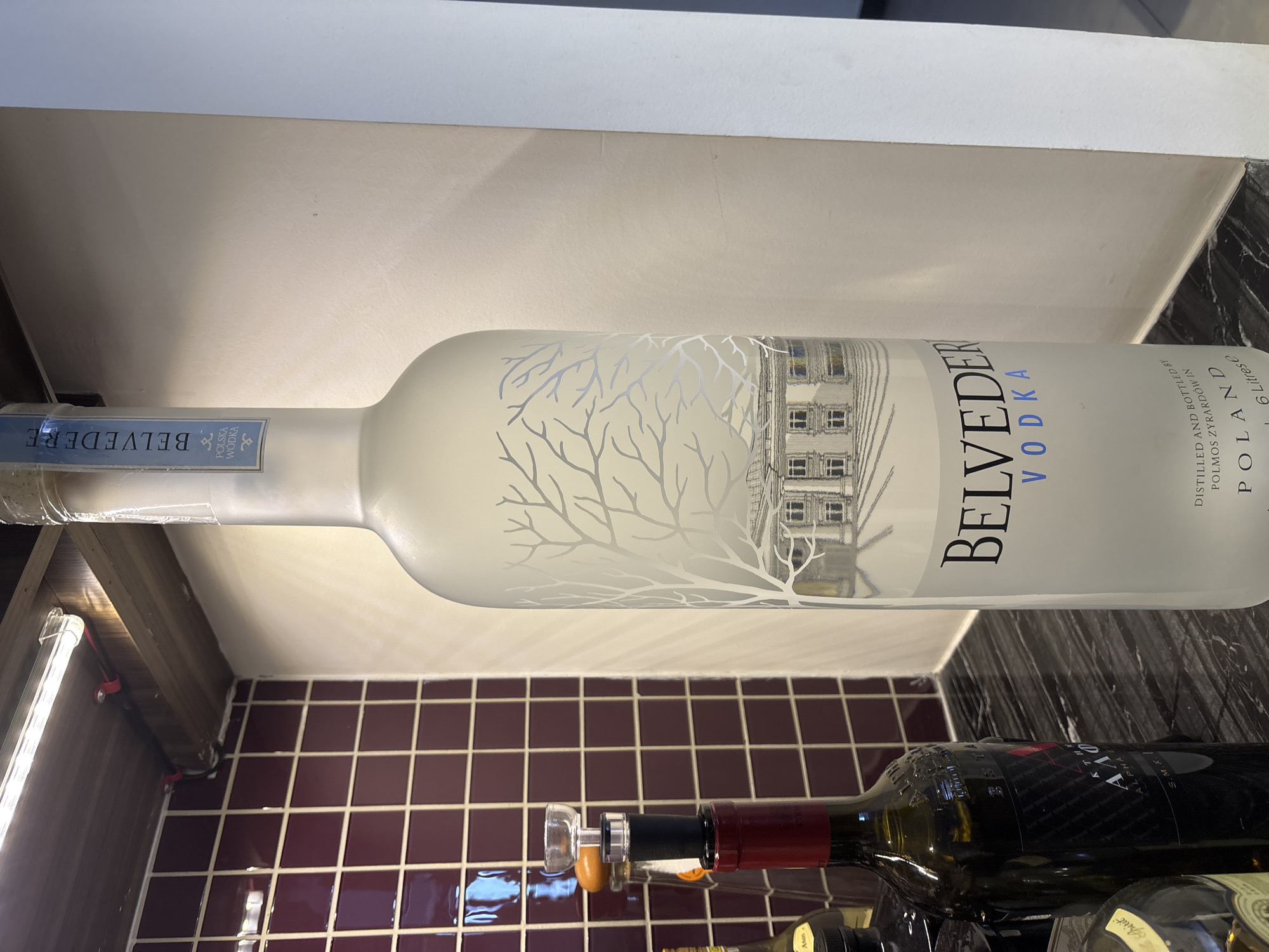The image shows a large bottle of Belvedere vodka standing on a marble countertop. Behind the vodka bottle, a dark red wine bottle with a black label is visible, topped with a wine stopper. The background features a tiled wall with a geometric pattern and a fluorescent light fixture mounted on a wooden surface. The scene is indoors, suggesting a domestic or commercial setting. There is no indication of the city of Dubai or the United Arab Emirates.