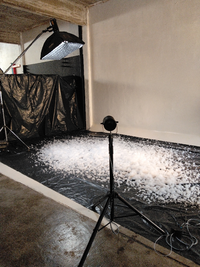A makeshift photography studio features a white wall backdrop and a black tarp covering the floor. Piles of white foam cover the tarp's surface. Professional lighting equipment includes a large softbox on a boom arm and a smaller spotlight on a tripod. Cables and equipment are scattered near the base of the spotlight tripod, suggesting a cluttered, functional workspace.