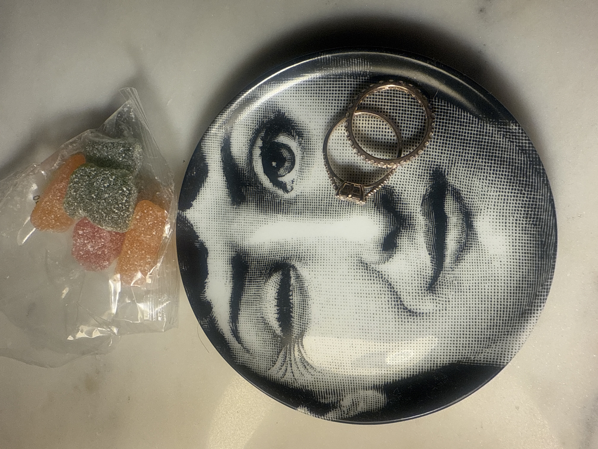 The image shows a still life composition on a white marble surface. A plastic bag filled with assorted colored and sugar-coated jelly candies is on the left. To the right, there is a black and white circular plate with a stylized face design. Two gold rings rest on the surface of the plate. Location information for Vila Nova de Gaia, Portugal, cannot be determined from this image.