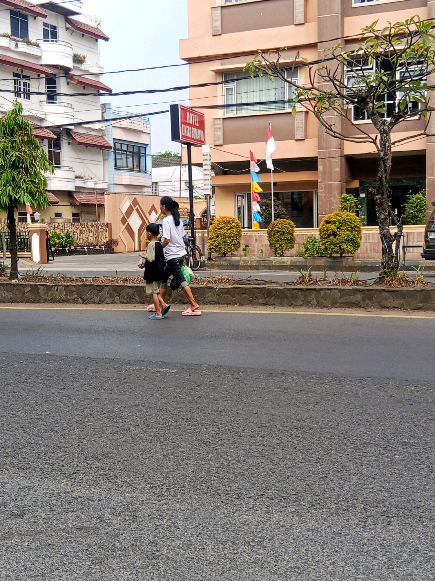 The image shows a street scene in the city of Lubuklinggau, Indonesia. Two people, an adult female and a child, are walking across the street. The adult female is wearing a white t-shirt and black pants, and the child is wearing a black t-shirt and beige shorts. They are walking from right to left. In the background, there are buildings, including a hotel with a red sign that reads "HOTEL LINTAS SUMATRA." There are also trees and bushes, and flags are visible on a flagpole. The street is paved with asphalt, and there is a stone wall along the sidewalk. The sky is overcast.