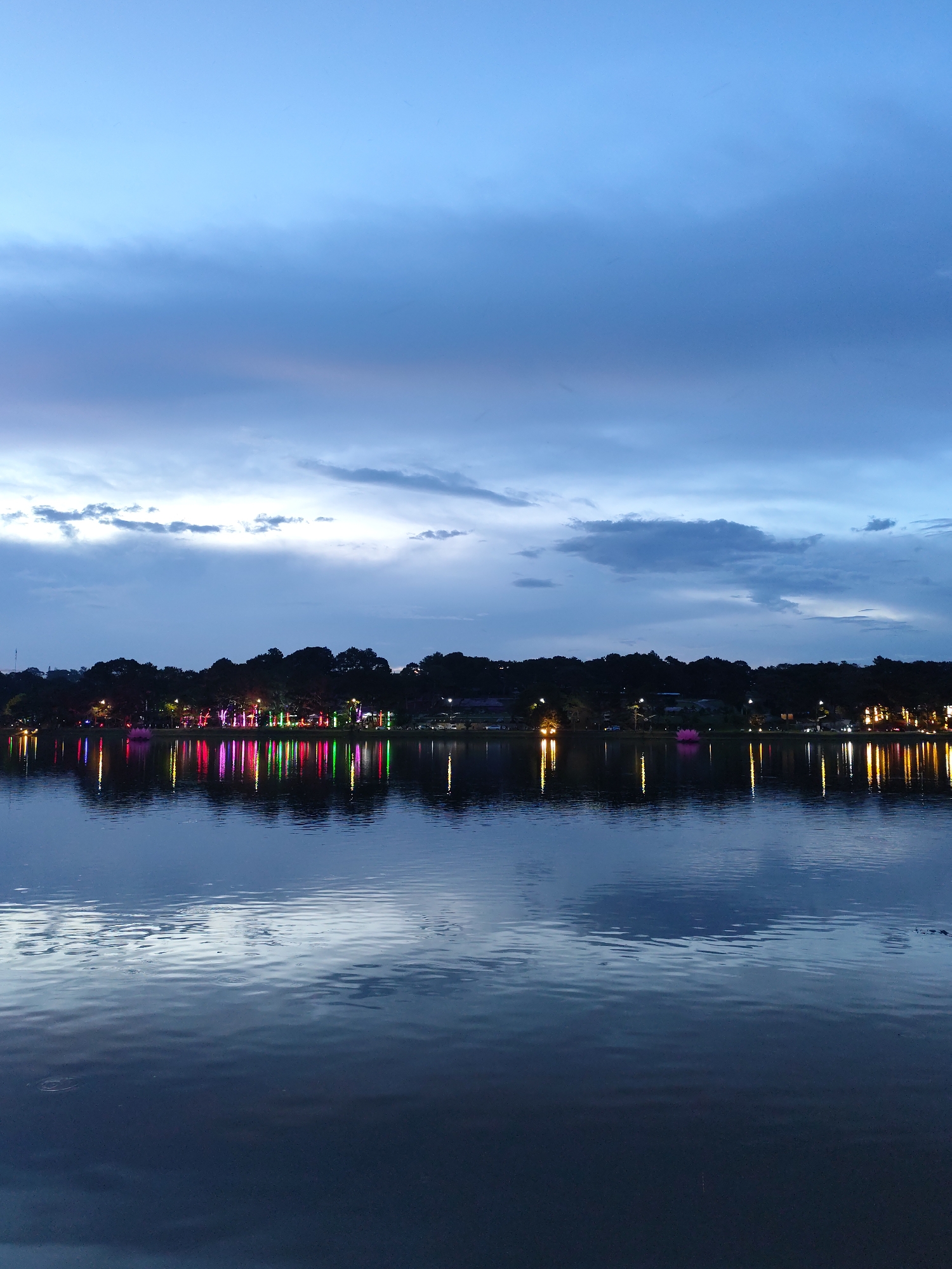 A nighttime view of a lake reflecting illuminated trees and buildings in the city of Unknown, Unknown. The sky is a mix of blue and gray clouds. The water is dark with visible reflections of lights from the far shore. There is no clear indication of any people, actions, or interactions within the frame. The environment appears tranquil and serene.