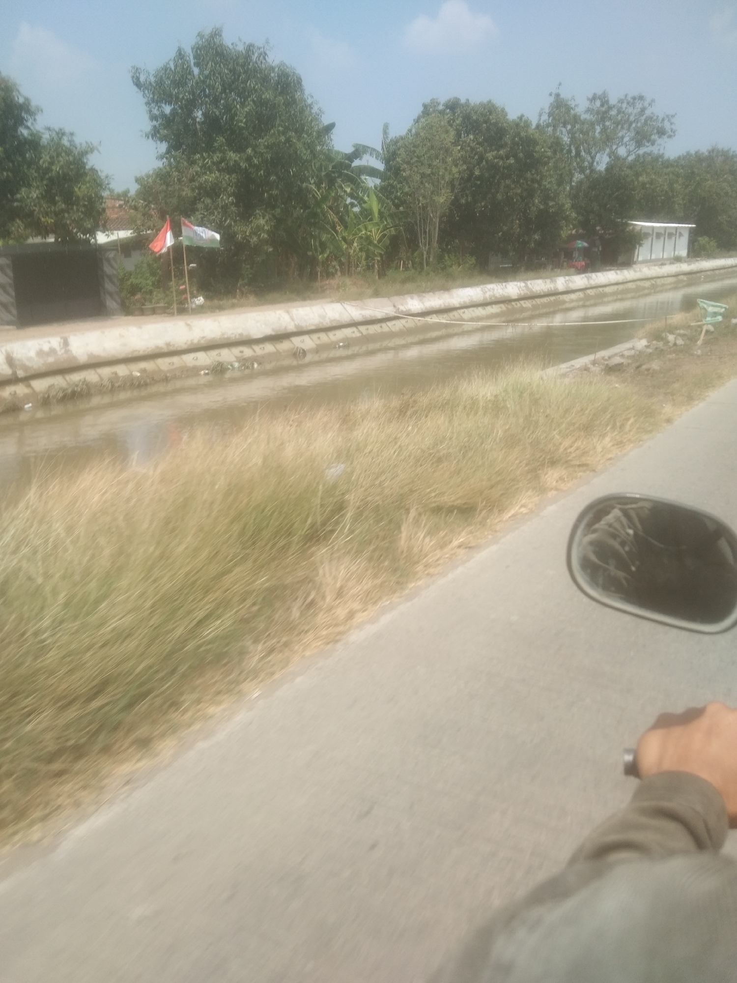 The image depicts a canal with concrete retaining walls, overgrown with dry grass on the right bank. On the left bank, there are trees, a building with a grey wall, and two Indonesian flags planted in the ground. In the distance, a white building with a blue roof is visible, along with more trees and vegetation. The scene appears to be in a rural or semi-rural area. The foreground shows a portion of a road and a motorcycle's side mirror. A person's hand and arm are visible, holding onto the motorcycle's handlebar. The sky is clear with some clouds. The location is identified as Desa Cilandak Lor, Indonesia.