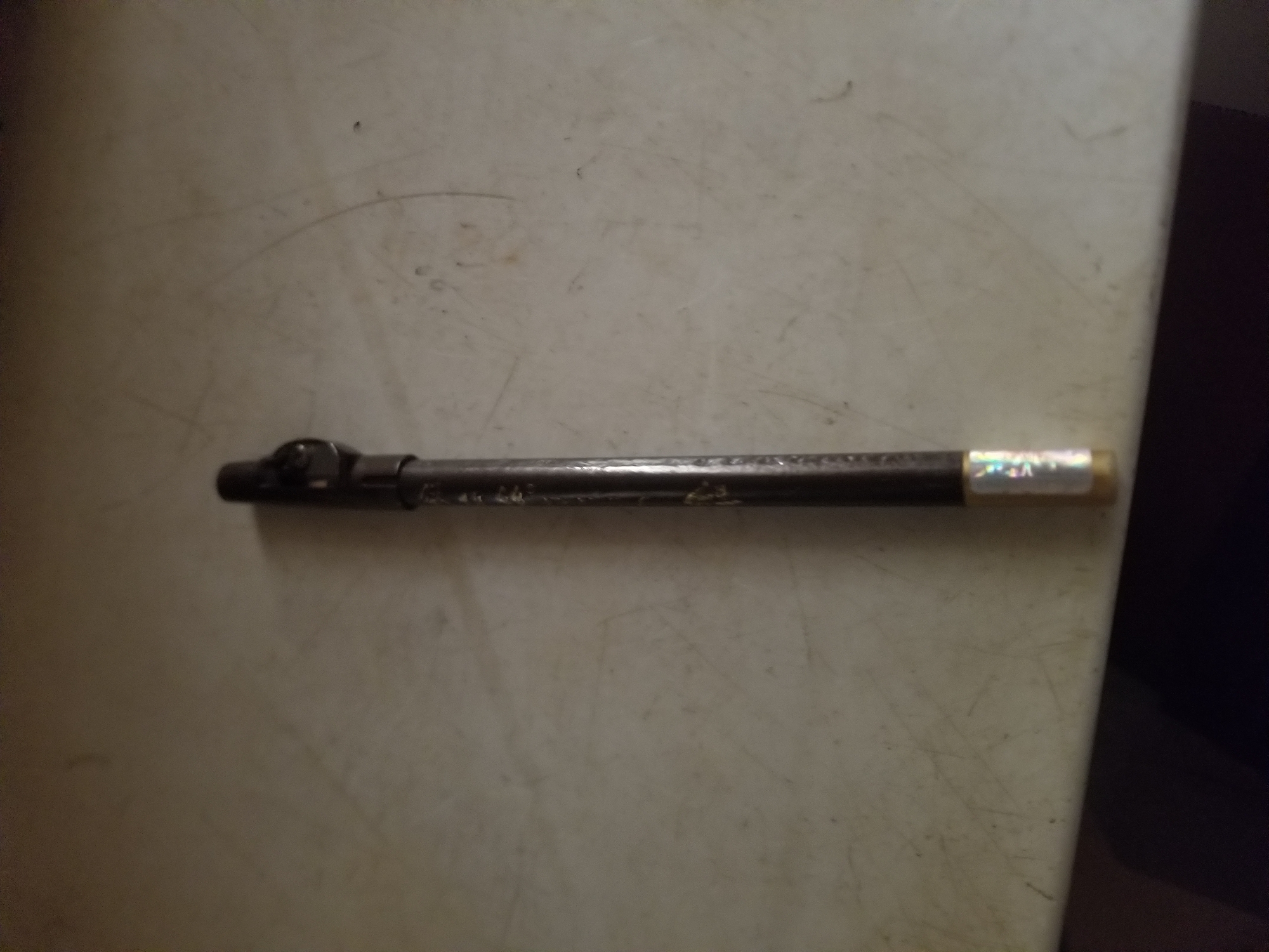 This media file shows a close-up, horizontal shot of a metal object lying on a textured, off-white surface. The object is dark and cylindrical, resembling a pen or a small tool, with some intricate metalwork at the left end. There is a small, rectangular label with a holographic or iridescent quality attached to the right end of the object. The surface it rests on appears to be a table or countertop, with visible scuff marks and faint curved lines. The lighting is dim, creating shadows and obscuring finer details of the background, which appears to be dark and out of focus on the right side of the frame. No people are visible, and the activity or event is not discernible beyond the object being placed on a surface. There is no discernible text on the object itself, although the label might contain some, it is illegible.