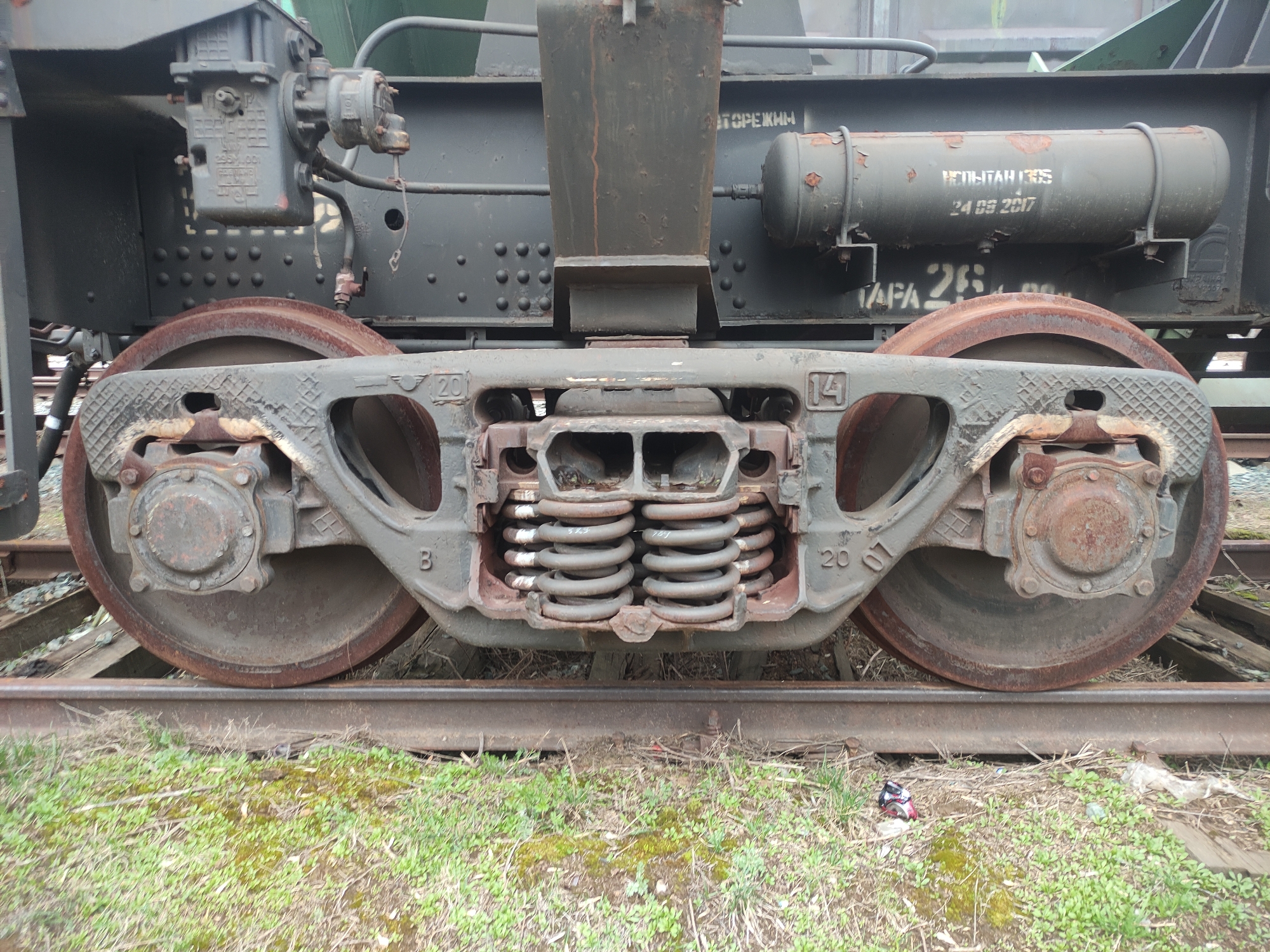 The image captures a low-angle view of the undercarriage of a stationary train car, specifically focusing on its bogie and two large, rusty metal wheels resting on railway tracks. The scene is set outdoors, likely in a rail yard or industrial area, with green grass and some visible debris (a crushed can and plastic bag) on the ground next to the tracks.
The train car's undercarriage consists of a complex assembly of weathered metal components. The prominent bogie frame, painted a dark grey, shows signs of wear and minor rust. In the center, two sets of heavy-duty coil springs are visible, designed to absorb shocks. The wheels are heavily rusted on their treads and inner faces, suggesting either age, heavy use, or disuse. Bearing housings are visible on the wheel axles, also showing rust. Above the bogie, part of the train car's body, painted a dark, muted green or grey, is visible, featuring numerous rivets and some attached equipment, including pipes and a cylindrical air tank.
The lighting is diffused, suggesting an overcast day. No human activity or specific event is taking place.
Several pieces of text are visible on the train components:
- On the cylindrical tank: "ИСПЫТАН ГУС" (Tested GUS) and the date "24 09 2017" (September 24, 2017).
- On the bogie frame, numbers like "20" and "14" appear multiple times, along with the letter "B".
- On the main body of the train car, partially visible text reads "ТАРА 26..." (Tare weight 26...).
- On a mechanical component attached to the upper left, faint Cyrillic text and numbers "295.001" are discernable.
- A word "ТОРЕЖИН" (TOREZHN) is written on a horizontal beam above the springs.