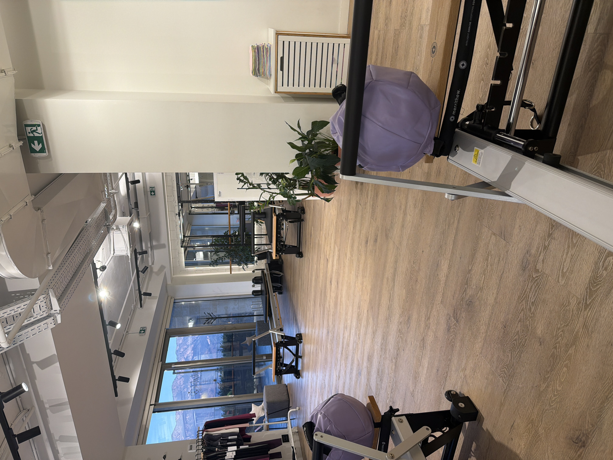 The image depicts the interior of a Pilates studio located in Boreti, Montenegro. The studio features a spacious, open-plan layout with wooden flooring and large windows offering a view of mountains and a body of water.
Several Pilates reformers are positioned throughout the room. In the foreground, a silver and black reformer is visible with a purple cushioned apparatus on it. Another reformer is seen further back in the center of the room, also with a purple cushioned piece. To the left, a rack of athletic wear is displayed.
The studio is well-lit with track lighting installed on the ceiling. Several potted plants are placed around the room, adding a touch of greenery. A white cabinet with slatted doors stands against a wall, topped with a stack of colorful folded items. An exit sign is visible on the ceiling.
The environment appears clean, organized, and ready for use. There are no people visible in the image. The overall impression is one of a modern and well-equipped fitness space.