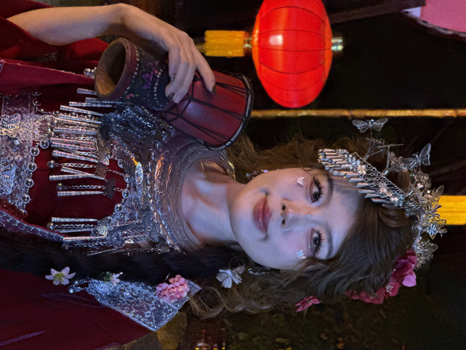 A female subject, appearing to be of East Asian descent, is depicted from the upper chest level, looking directly forward in a nighttime setting. She wears an elaborate silver metallic headdress featuring butterfly motifs and various dangling ornaments, along with pink and white floral adornments in her styled brown hair. Her face is made up with prominent eyelashes, light-colored contact lenses, pink lipstick, and small, reflective butterfly appliques on her cheeks. She also wears large, dangling earrings.
Her attire consists of a red garment, layered with a prominent silver metallic necklace or chest piece. This piece covers her upper chest and shoulders, displaying intricate patterns, fan-shaped elements, dangling cylindrical pieces, and small bells. An outer layer of red velvet-like fabric is visible, with silver embroidered trim on what appears to be a lapel or collar.
In her left hand, she holds a small, reddish-brown drum, possibly a djembe, with black lacing and decorative patterns near its base. The drum rests against her left shoulder area.
The background is dark, indicating a nighttime environment. To the left and slightly behind the subject, a large, illuminated red lantern with a yellow tassel hangs. Additional blurred, warm-toned lights are visible in the distant background, consistent with an urban or festive area in Yuzhong District, China.