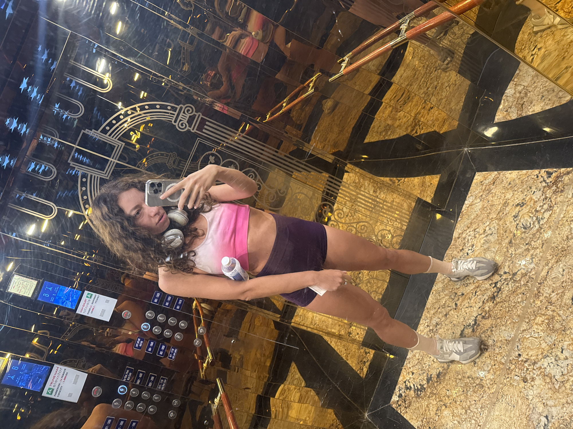 A full-length, eye-level shot depicts a woman taking a selfie in an elevator. She is holding a cell phone in her left hand and a water bottle in her right. She is wearing a pink and white sports bra, purple shorts, white socks, and gray athletic shoes. She has white headphones around her neck. She is standing on a patterned floor and the elevator walls are shiny and reflective. The control panel of the elevator is visible in the background. A sign reads "TRUNG NAM elevator". The text on the elevator's control panel identifies the city of Hoi An Ward, Vietnam.