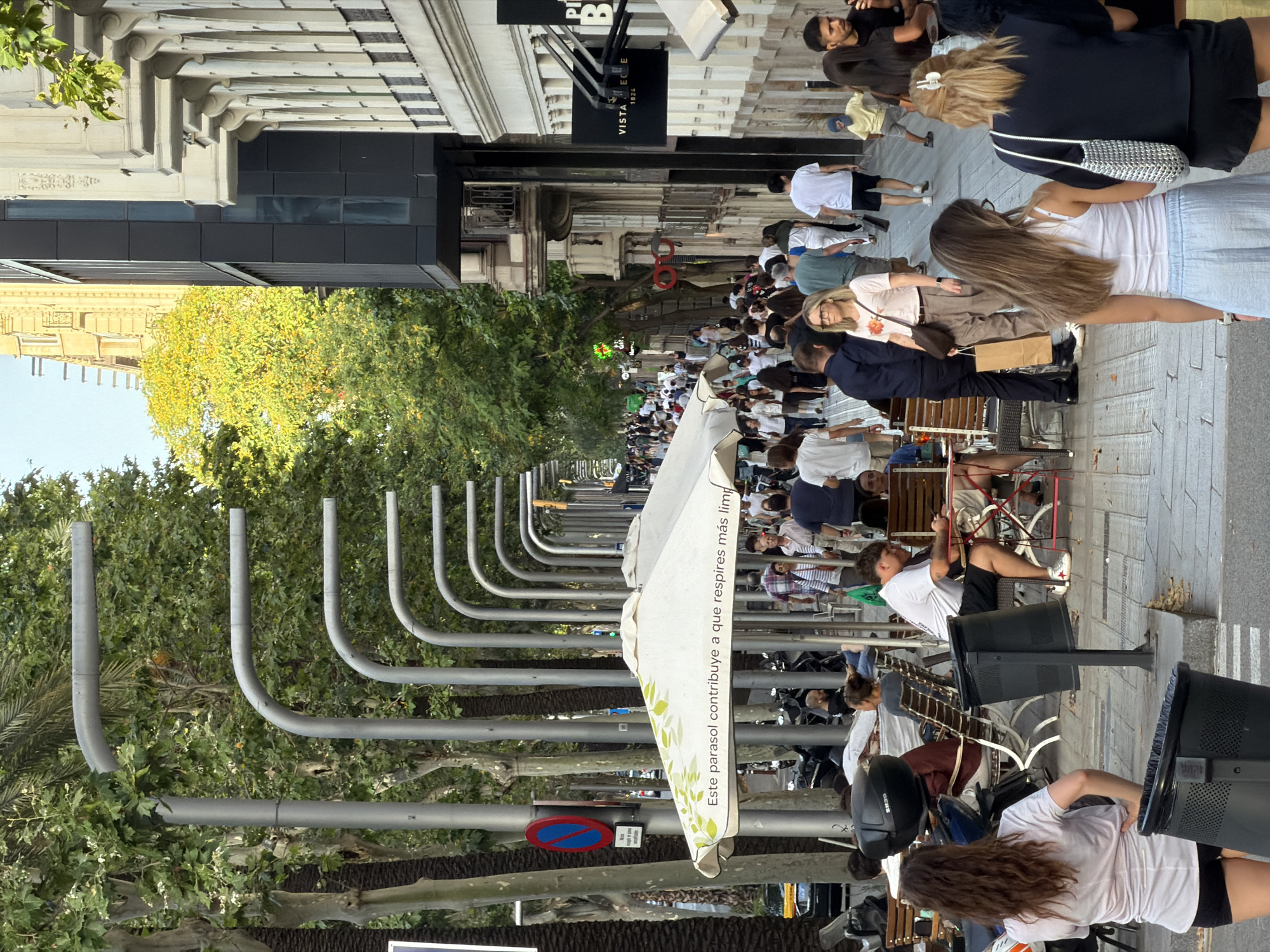 This is a street scene in Barcelona, Spain, likely on a pedestrianized boulevard. Tall trees line both sides of the street, with palm trees and deciduous trees visible. Numerous street lamps with curved arms are installed along the pathway. Buildings with ornate facades are visible on the right side of the image, with modern architectural elements also present. A large white umbrella with a green leaf pattern and text that reads "Este parasol contribuye a que respires más lim" is prominent in the foreground, suggesting it's part of an outdoor seating area for a cafe or restaurant.
A large number of people are walking along the wide pedestrian sidewalk, creating a lively atmosphere. Many are dressed in casual summer attire. Some individuals are seated at tables, also likely associated with outdoor dining. Several motorcycles are parked along the left side of the street, beyond the seating area. A blue and red circular sign with a diagonal bar is affixed to a pole on the left, indicating a no-parking or restricted access zone. The ground is paved with grey interlocking stones. The time of day appears to be late afternoon or early evening, given the warm sunlight and shadows.