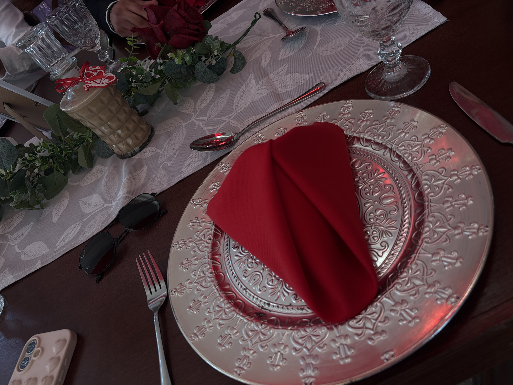 The image shows a table setting, possibly in a restaurant or dining room, with an ornate silver charger plate topped with a folded red napkin. A fork is placed to the left of the plate, and a knife to the right. A spoon lies on the table near the top of the plate. A glass of water is visible in the upper right of the image, while another glass and a decorative bottle with a red ribbon and tag are on the table to the left. A pair of sunglasses and a mobile phone in a pink case are also on the table. The table is covered with a patterned runner.
There is no information within the image to pinpoint the specific location in Chilpancingo, Mexico.
