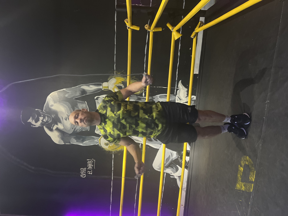 A man in a patterned shirt and black shorts stands in a boxing ring installation, his arms outstretched, gripping the yellow ropes. Behind him is a mural depicting a boxer. The scene is lit with a purple hue on the lower left. The floor is dark with a yellow letter "P" visible in the lower right. The perspective makes it appear as though the man is lying down.