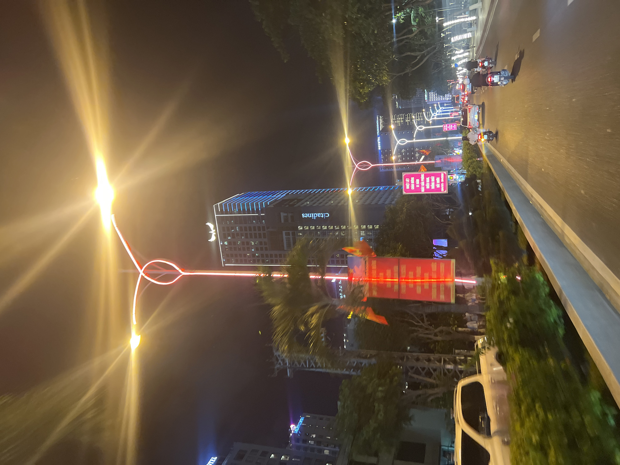 At night, the image shows the Citadines building lit with blue lights and a light post with intersecting red fluorescent shapes. A billboard with Vietnamese text stands nearby. The scene is on a street in the city of Nha Trang Ward, Vietnam. In the background, other illuminated buildings are visible, including a sign for Novotel. Vehicles, primarily motorbikes, travel on the road. Palm trees and other foliage line the street. The perspective is from a vehicle moving along the road.