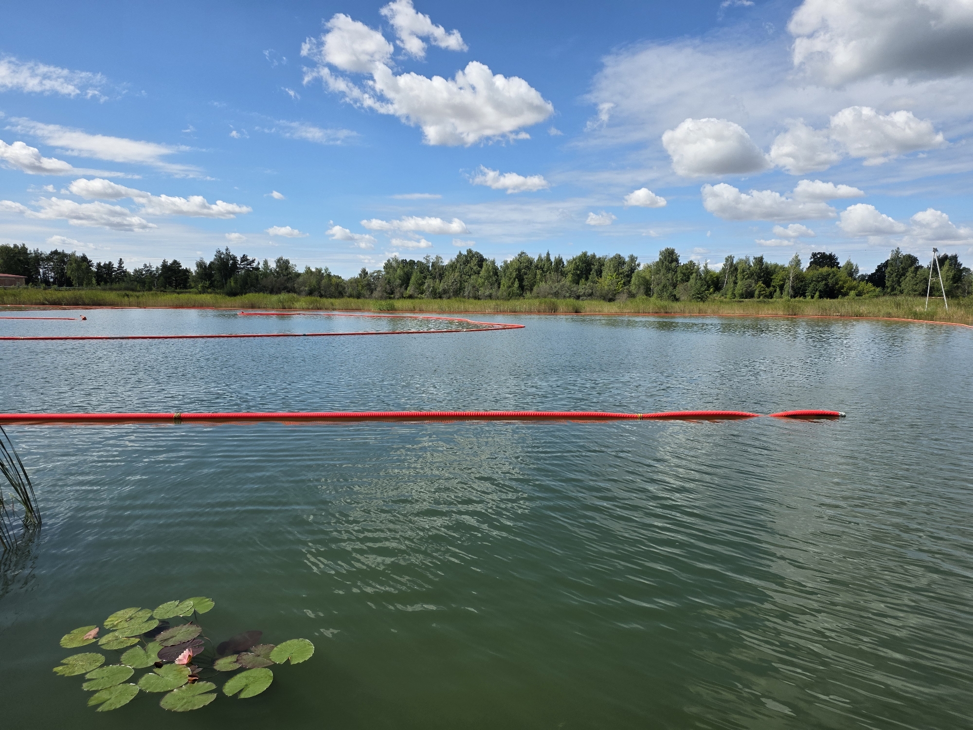 The image shows a body of water, likely a lake or pond, with a line of red buoys or floating barriers extending across the surface. In the foreground, several lily pads are visible on the water. The background features a dense line of trees and vegetation along the shoreline, under a blue sky with scattered white clouds. The scene appears to be an outdoor natural environment. There are no people visible in the image. The buoys suggest some form of containment or demarcation is in place on the water. The overall impression is one of a serene, possibly managed, aquatic environment.