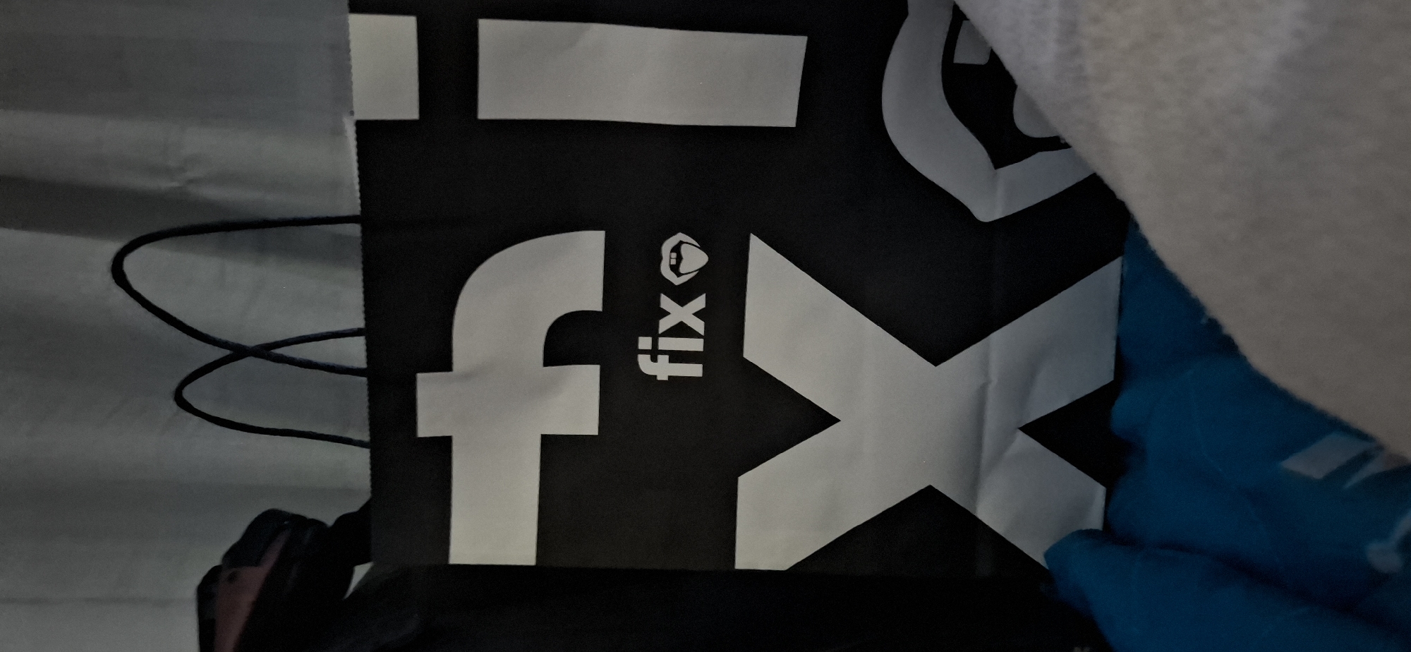 The image shows a black shopping bag with white lettering and graphics. The lettering appears to spell out "FIX" twice. The bag has two black handles. The background is out of focus and appears to be a light-colored fabric, possibly a curtain. The bag is lying on top of some blue and white fabric. There are no people visible in the image. The image does not provide any information about the location in the city of Yerocham, Israel.