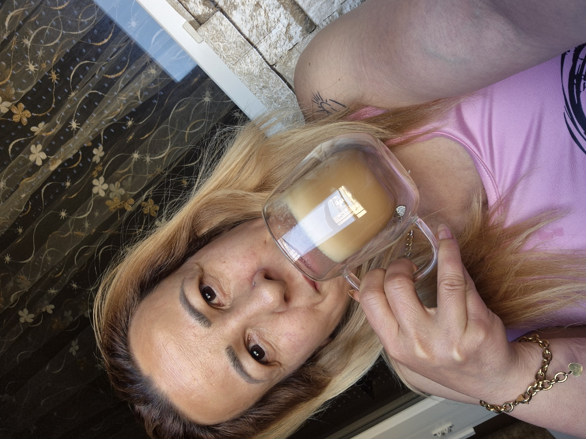 A woman with blonde hair and a pink top is taking a selfie while holding a clear glass mug filled with a light-brown beverage. She is wearing a gold bracelet with a heart charm. The background shows a textured wall and a curtain with a floral pattern.