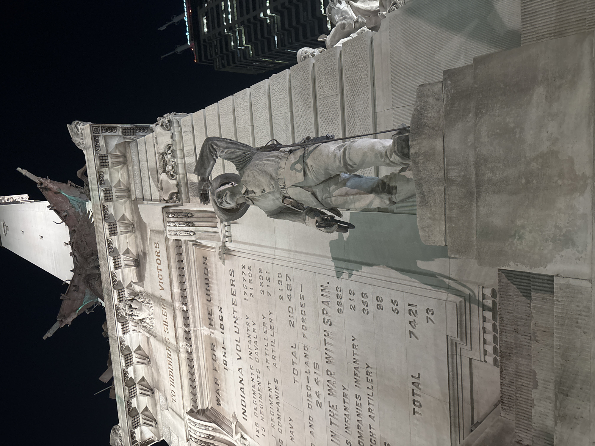 The image shows a portion of the Soldiers and Sailors Monument in Indianapolis, Indiana, United States. The monument, constructed of limestone, features inscribed text detailing Indiana's contributions to the Union during the American Civil War and the Spanish-American War. A life-sized bronze statue of a soldier in uniform is mounted on the monument, positioned with a weapon in hand. The monument includes elaborate architectural details such as ornate carvings and tiered stone blocks. The background shows buildings and the night sky.