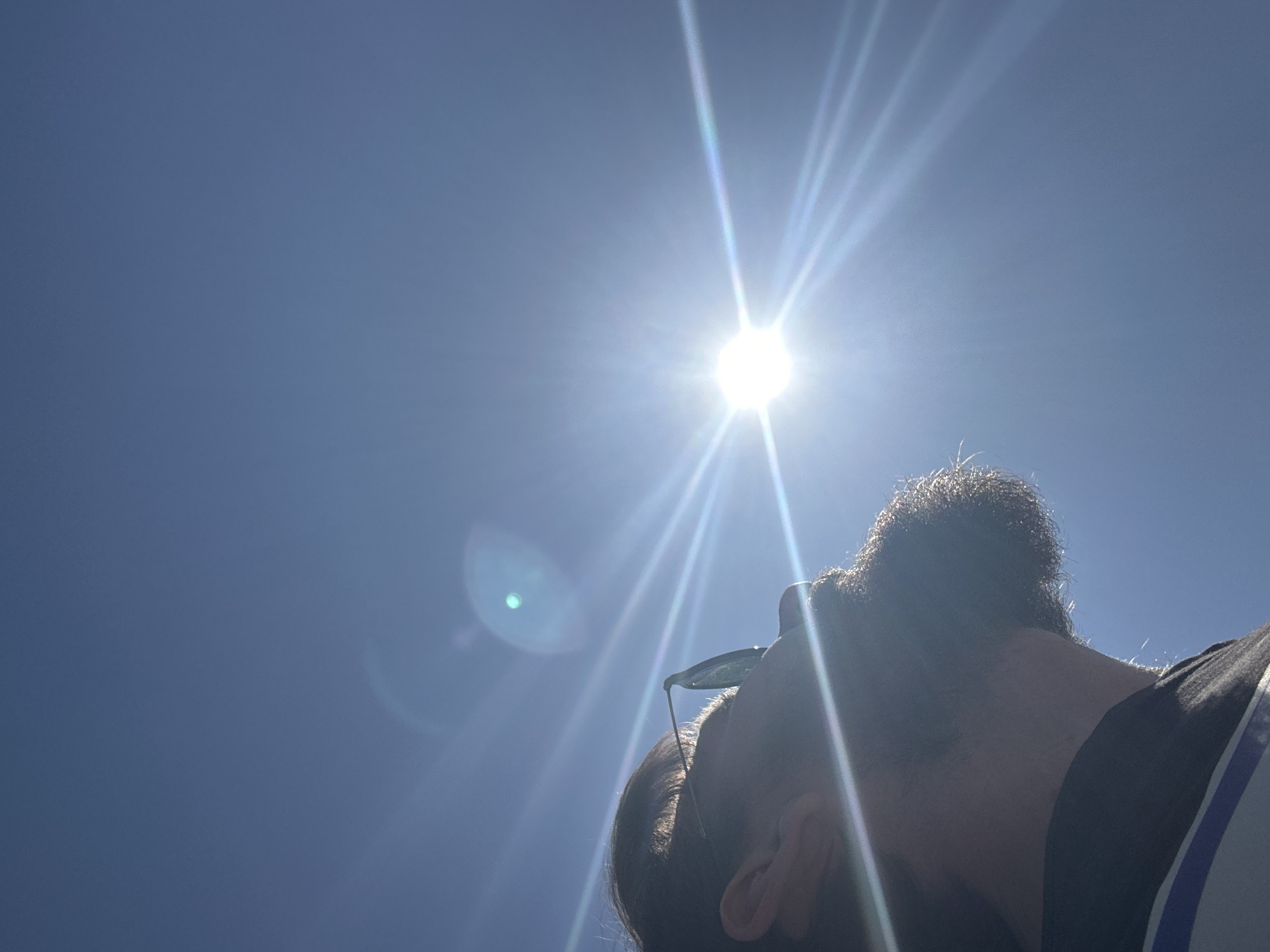 The image captures a low-angle view of a person looking up at the bright sun against a clear, blue sky in Fethiye, Turkey. The sun, positioned in the upper center, is intensely bright, emitting strong lens flares and starburst rays that spread across the sky. A circular, greenish-blue lens flare is also visible to the left of the sun.
The person's head and upper body are visible in the lower right portion of the frame, largely silhouetted against the powerful backlight from the sun. They have a beard and what appears to be dark hair styled in a bun or ponytail. A pair of sunglasses is perched either on their head or nose, with the frame visible. The person's face is turned upwards, directly towards the sun. The sky is a uniform, vivid blue with no clouds, suggesting a clear and sunny day. The overall impression is one of enjoying the bright sunshine and clear weather. There is no visible text in the image.